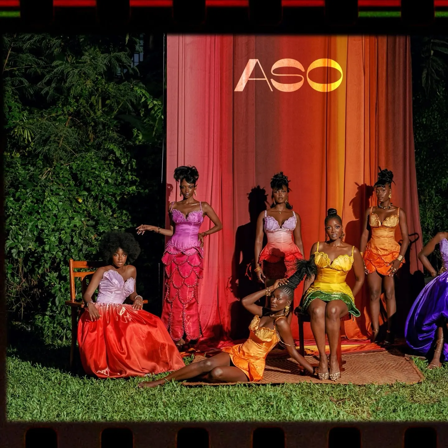 Seven models in sunset-hued satin gowns pose on a lawn before a red-orange curtain bearing the ASO logo.