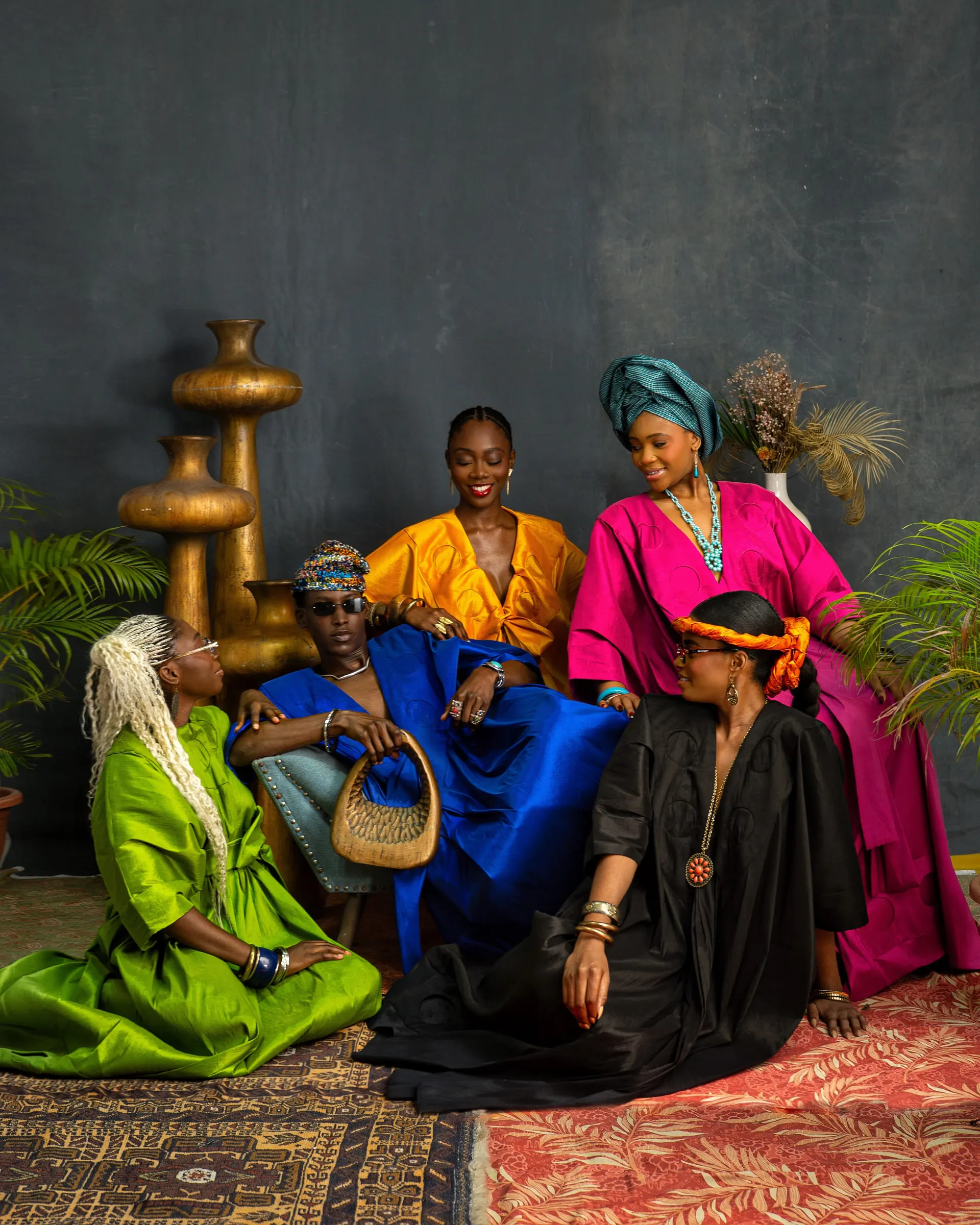 Five models in boubous in lime green, cobalt blue, marigold orange, magenta pink and black gather among tall gold vessels on a grey studio set, two seated on layered rugs and one in a pale armchair holding a woven bag.