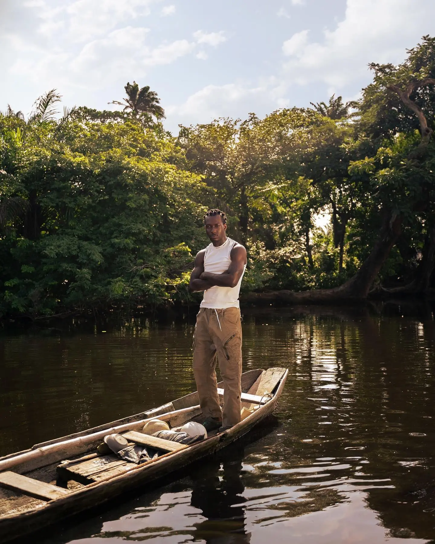 Savage Tha Beast stands arms crossed in a wooden canoe on a calm Niger Delta river.