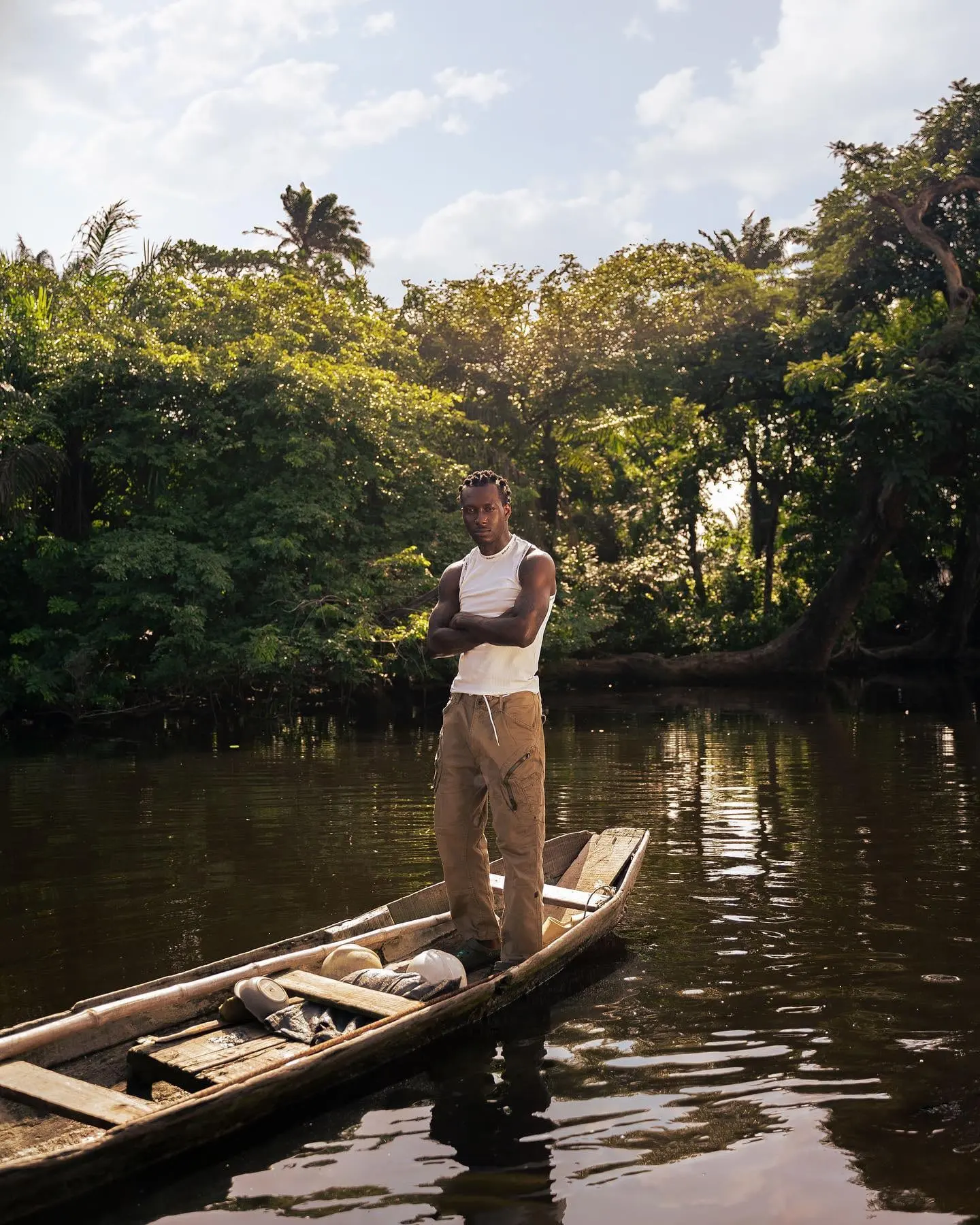 Savage Tha Beast stands arms crossed in a wooden canoe on a calm Niger Delta river.
