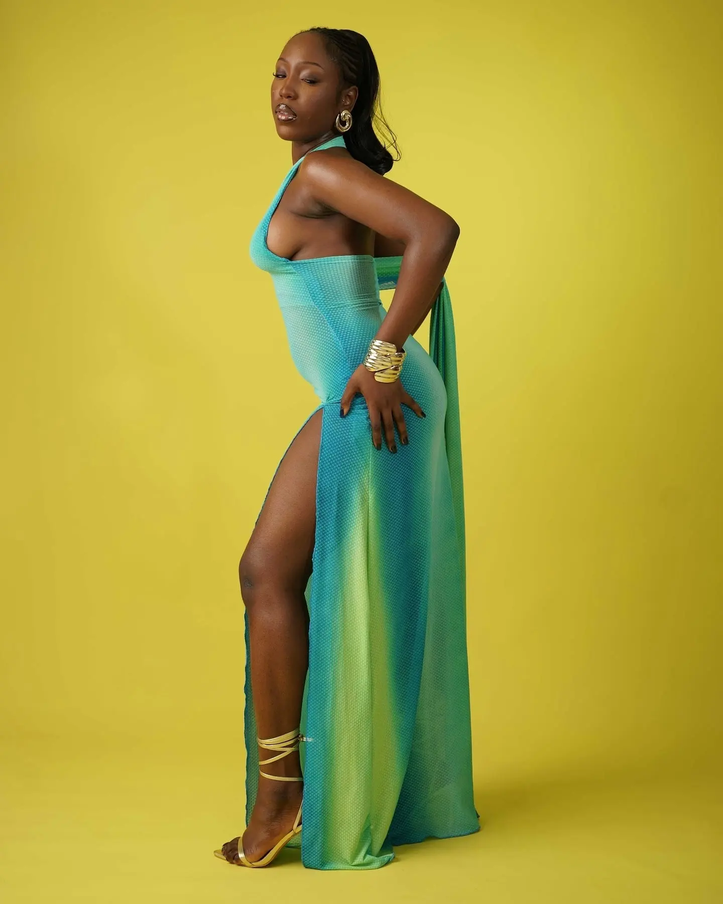 Model in profile wearing a turquoise-green halter gown with high slit and gold accessories on yellow.