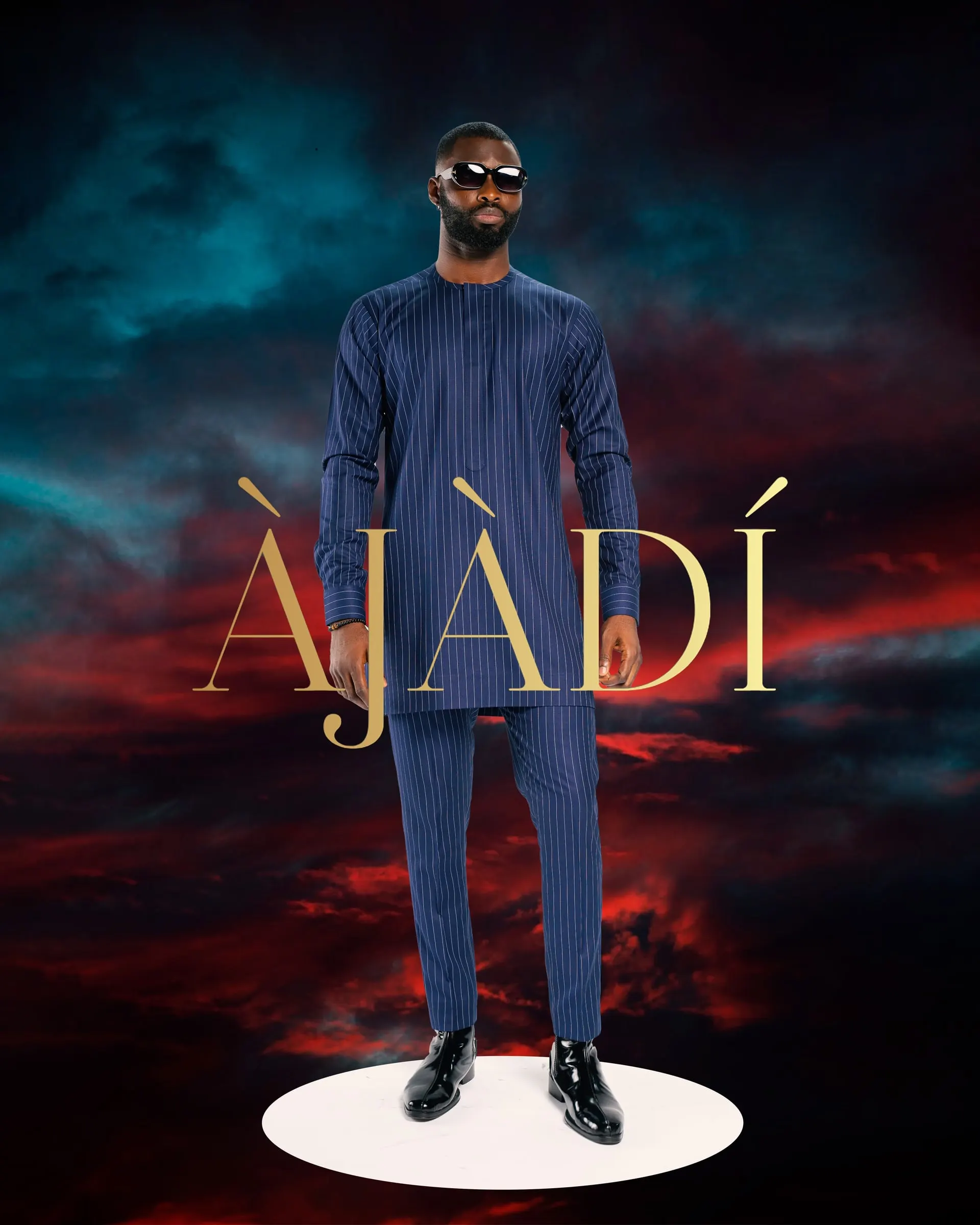 Model in sunglasses and a navy pinstripe tunic set stands on a white oval plinth against a red and teal storm sky, the gold ÀJÀDÍ wordmark beside him.