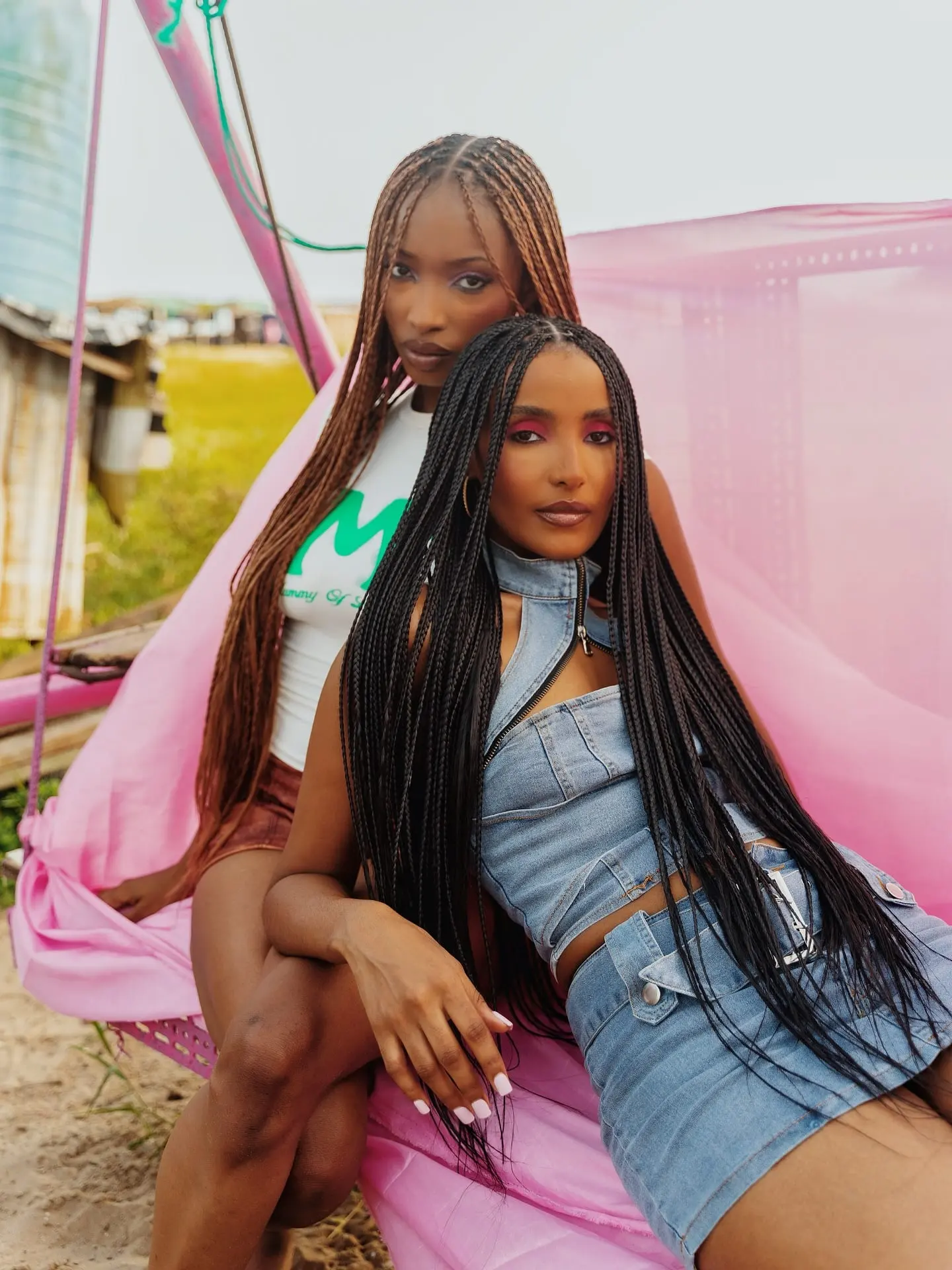 Two models with long knotless braids, one in double denim, sharing a pink fabric swing outdoors.