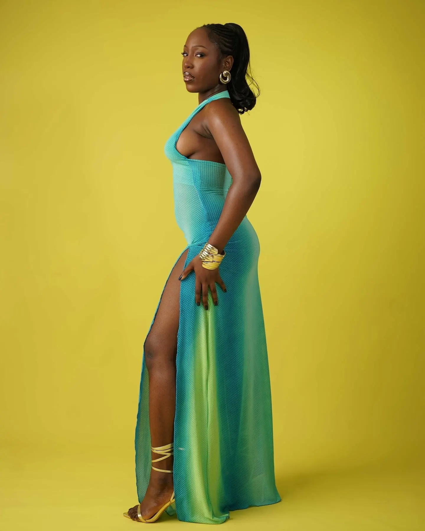 Model glancing over her shoulder in a backless turquoise ombré gown with gold cuff and strappy heels.