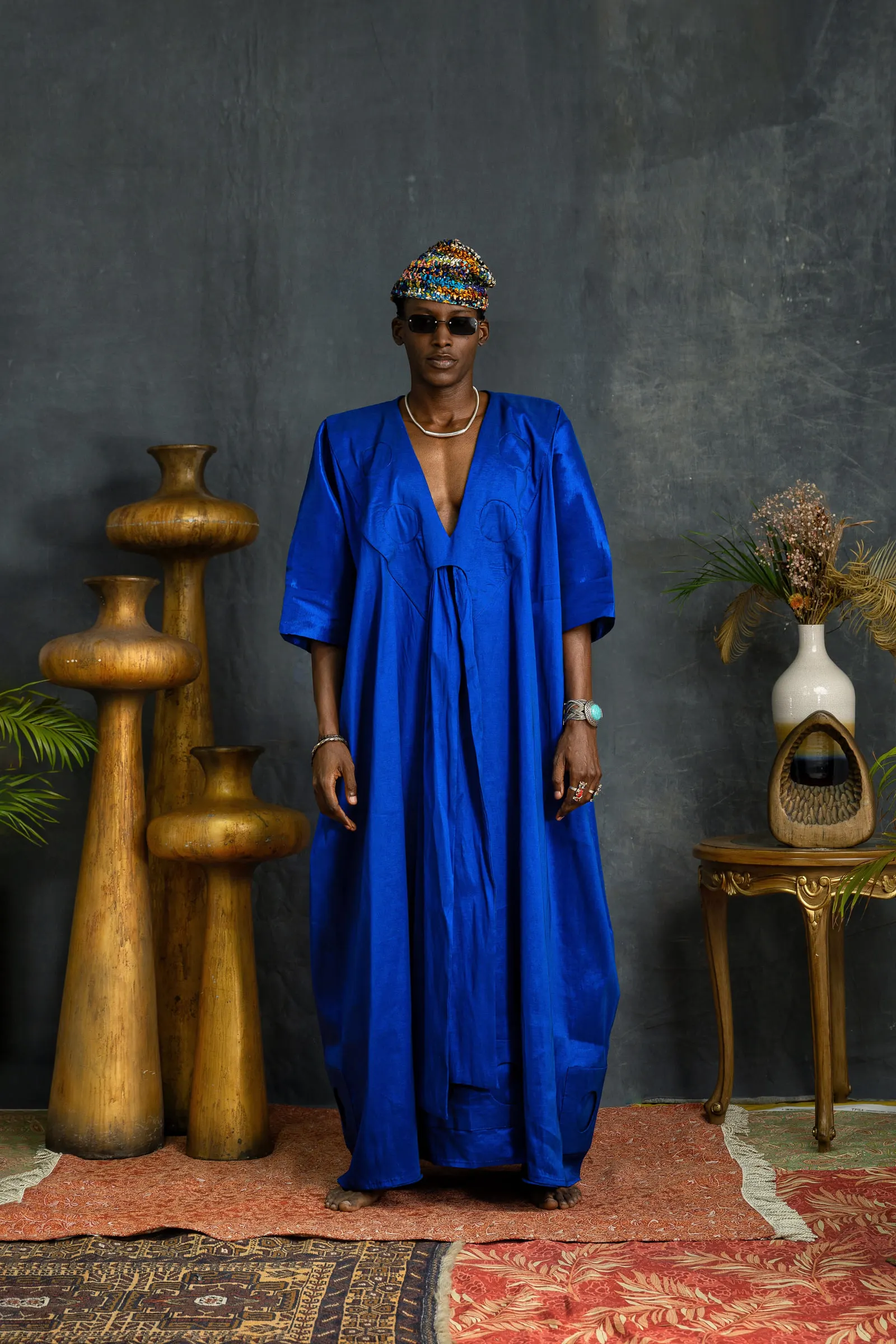 Model in a beaded cap and dark sunglasses stands in a cobalt-blue boubou between tall gold vessels, a gilt side table and a vase of dried grasses behind.