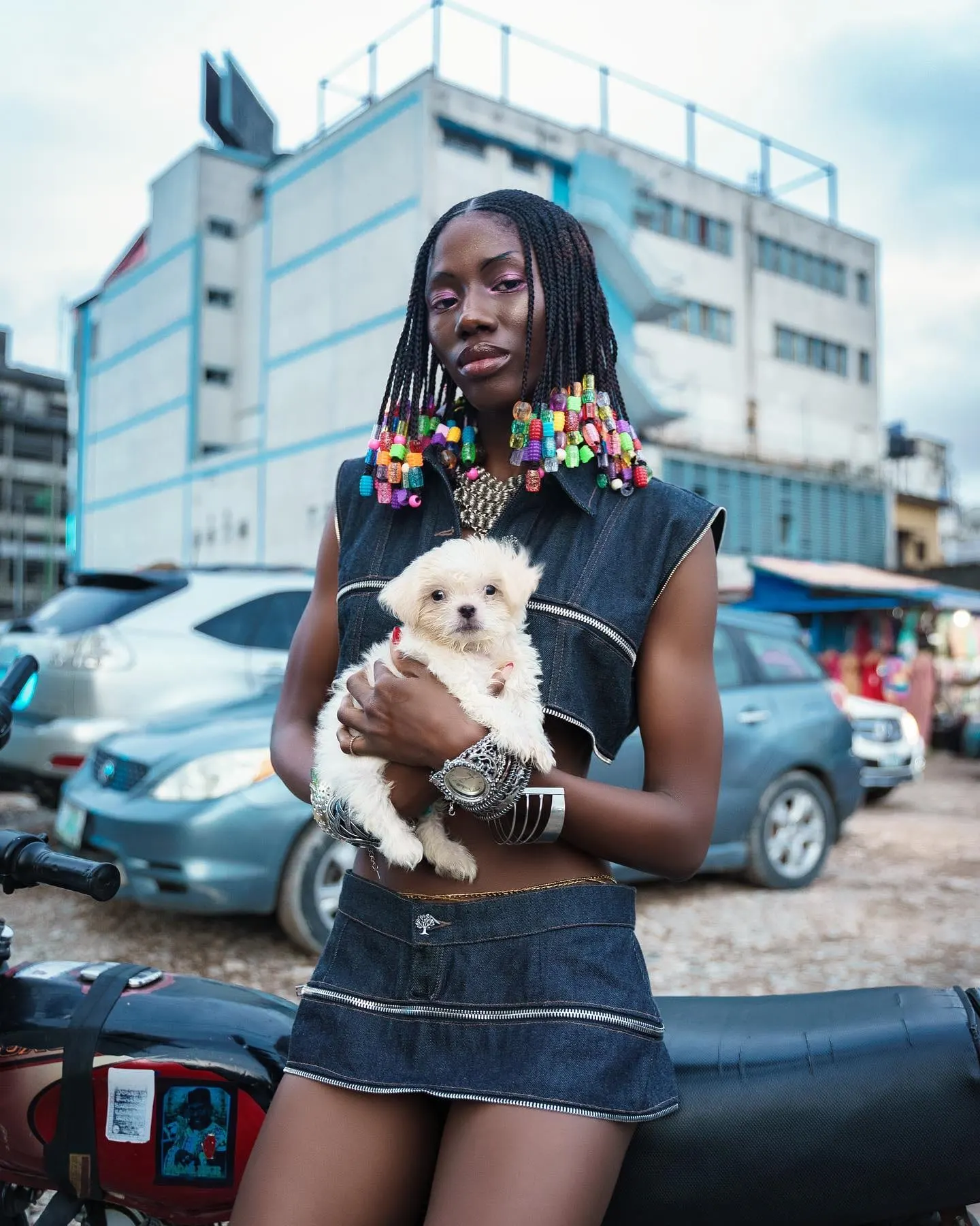 Model with beaded braids and a denim zip vest cradles a white puppy in a busy Lagos parking lot.