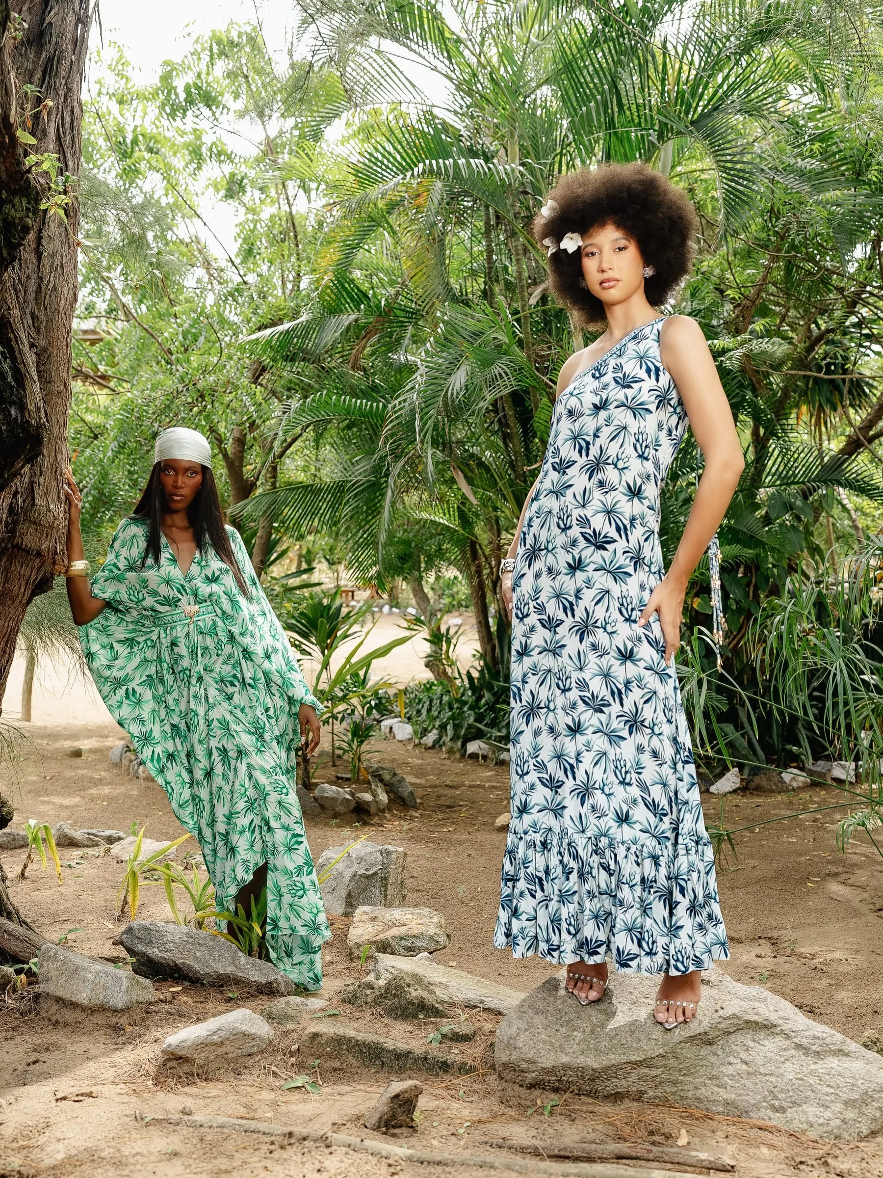 Two models among trees: one in a flowing green printed kaftan leans on a trunk, the other in a one-shoulder blue palm-print maxi dress stands on a rock with a white flower in her afro.