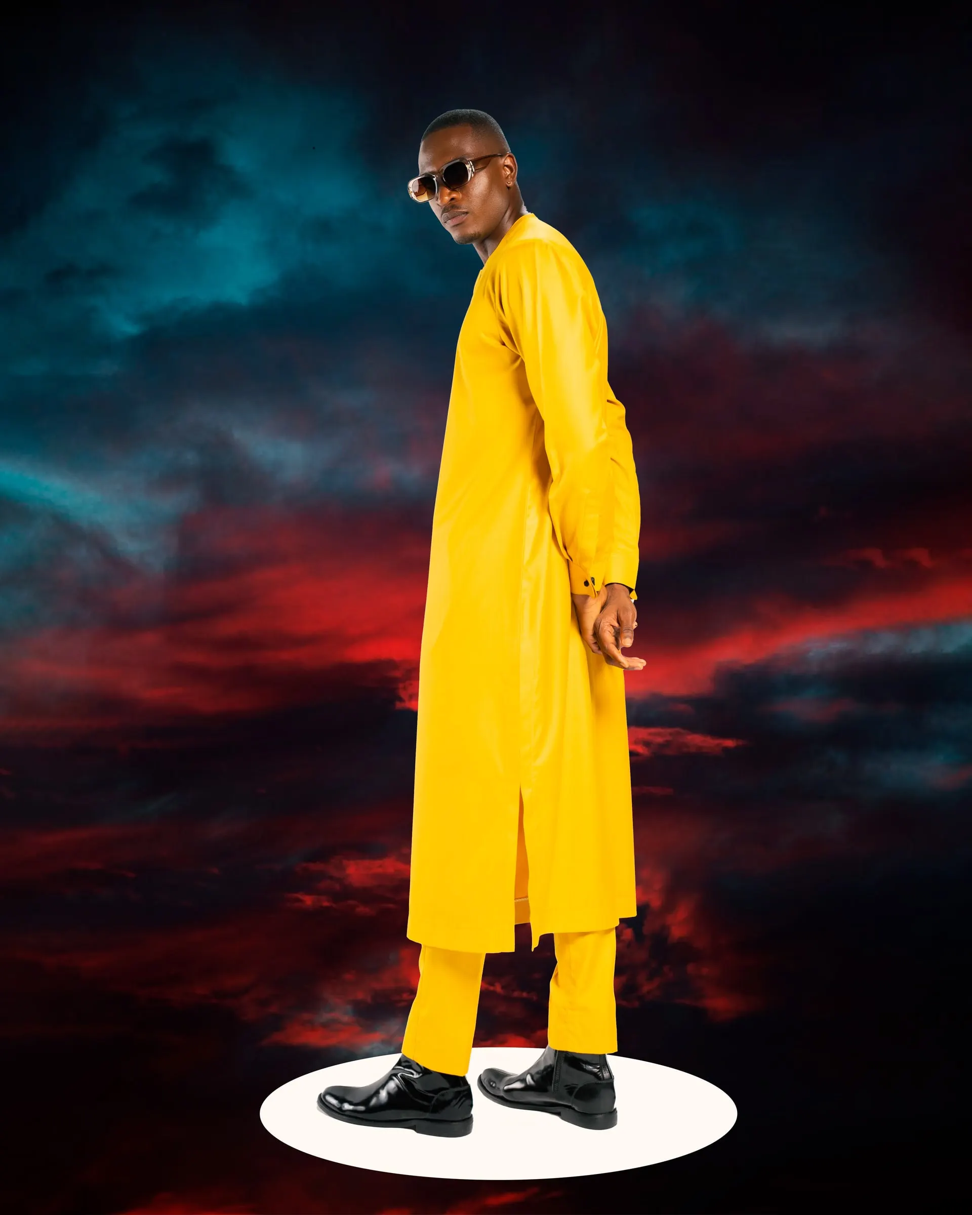 Model in a long yellow kaftan and matching trousers glances over his shoulder on a white plinth against a red and teal storm sky.