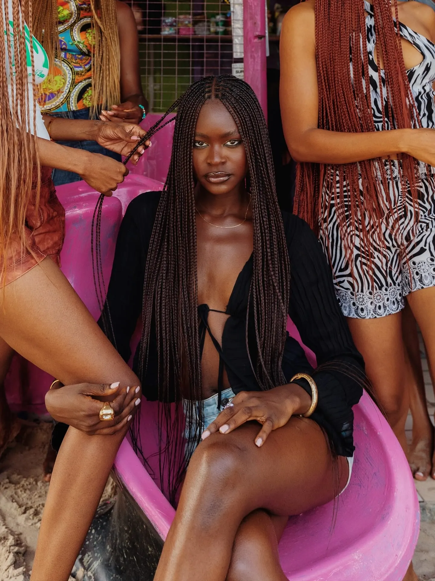 Model seated in a pink chair having her waist-length braids styled by hands of women around her.