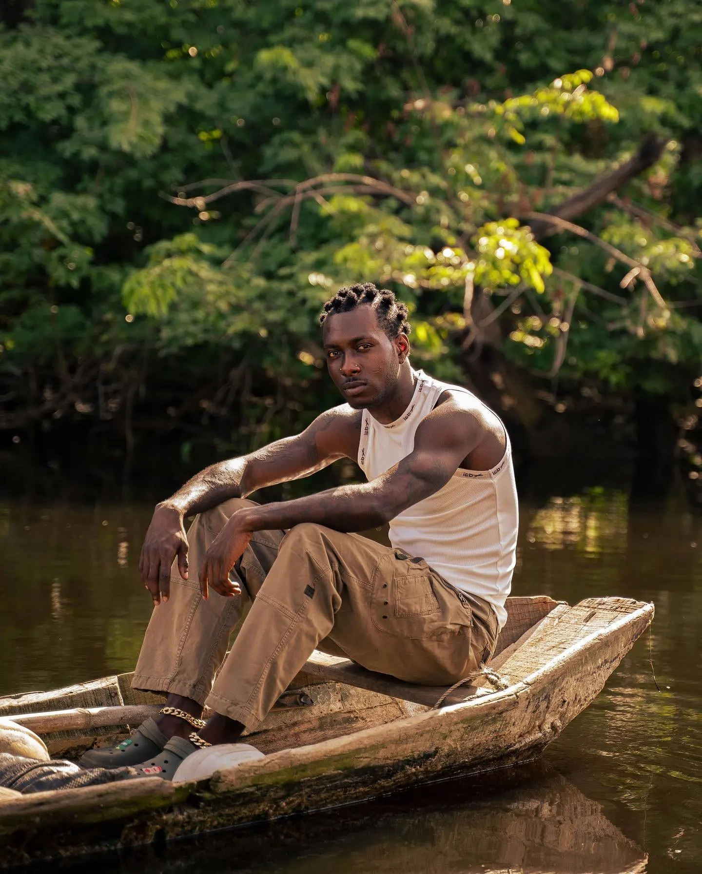 The artist, in white tank top and khaki cargos, sits on a dugout canoe's prow in golden light.