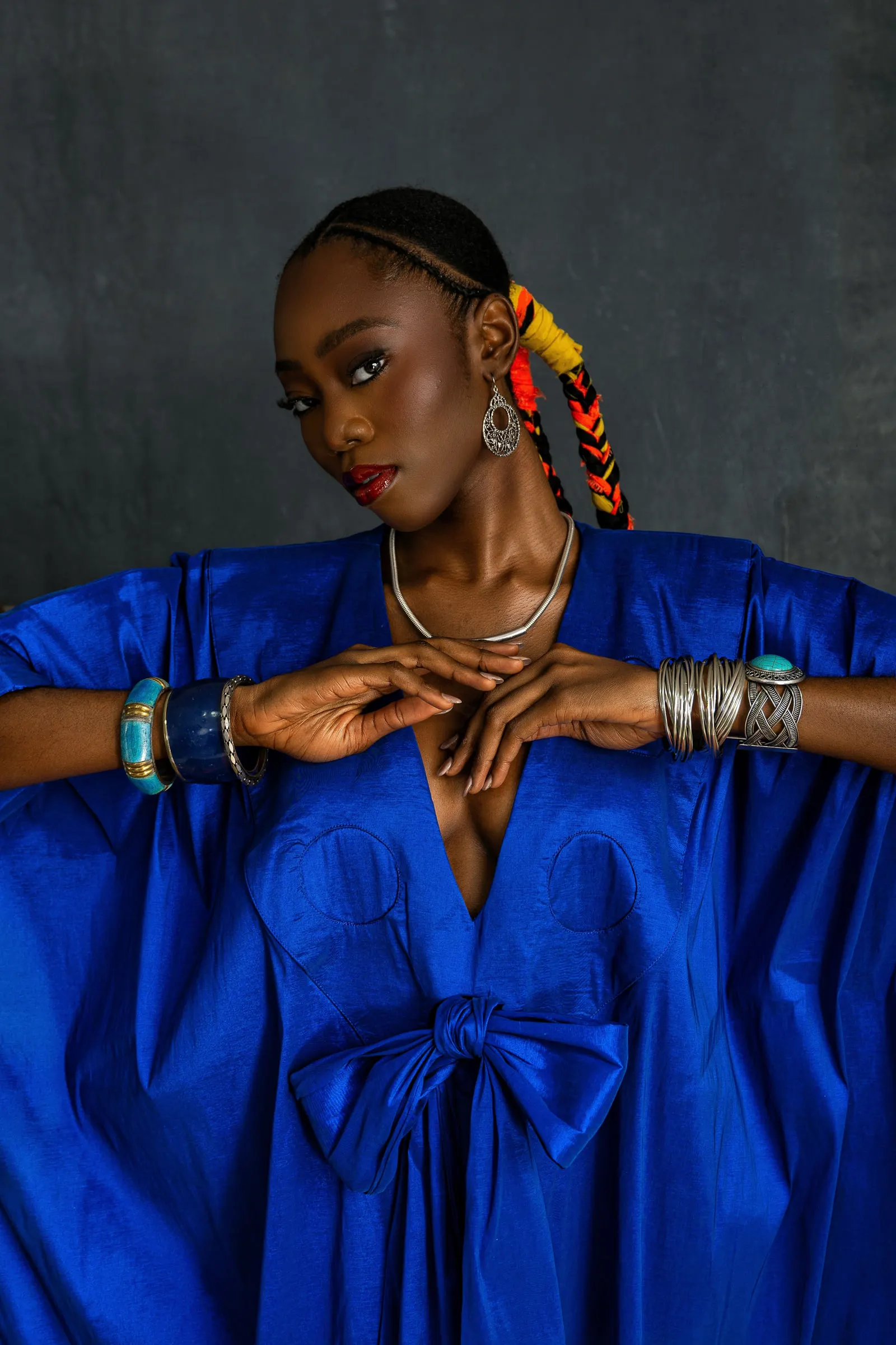 Close portrait of a model in a cobalt-blue boubou with a bow at the chest, wrists crossed at the collarbone under stacked turquoise and silver bracelets, an orange-and-yellow braid falling over one shoulder.