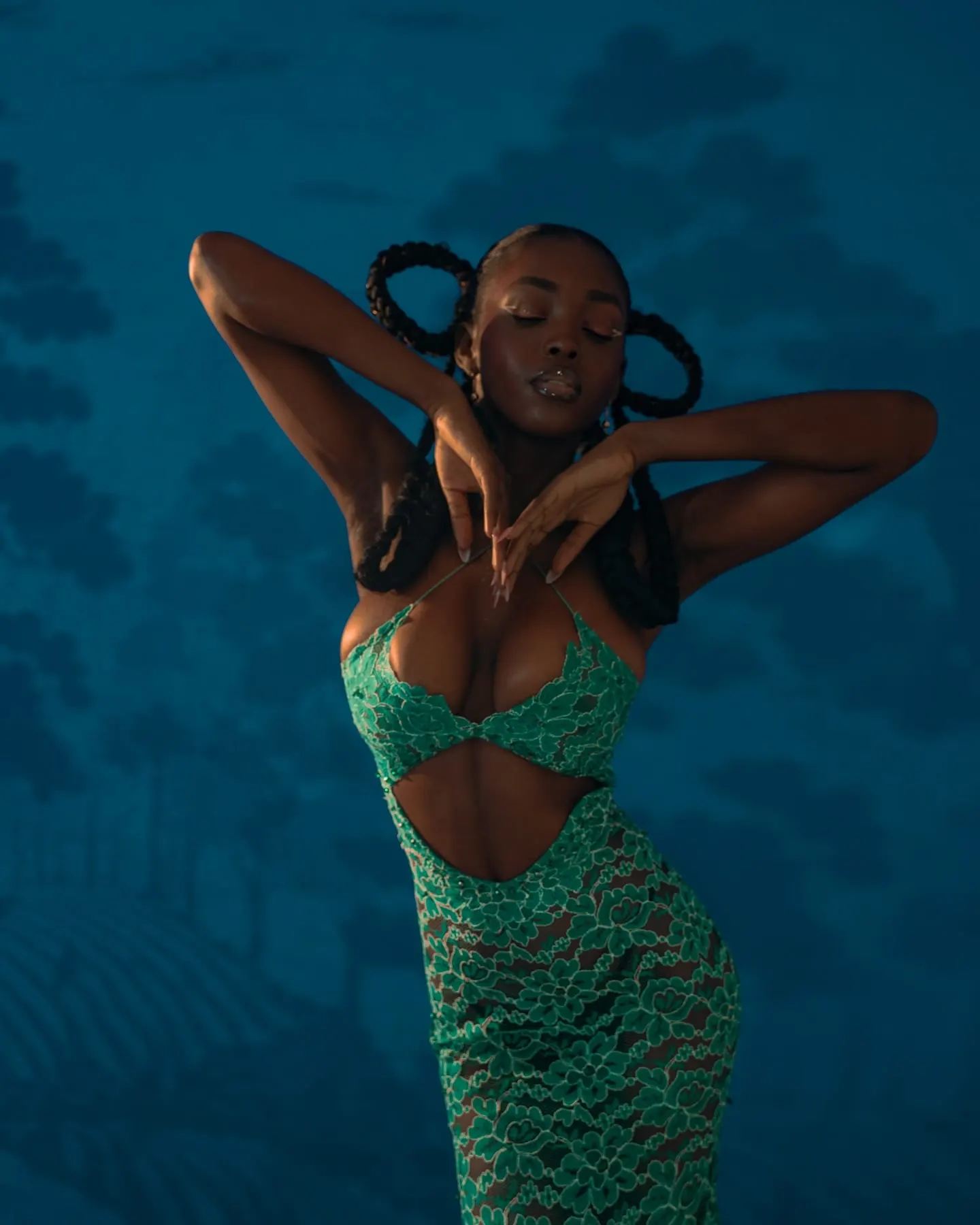 Uyi sways with eyes closed, wrists crossed at her chest, looped braids framing her face.