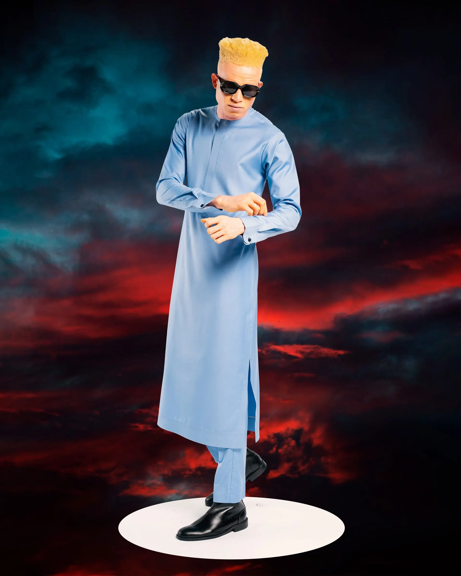 Model with a blond flat-top and sunglasses fastens the cuff of a long light-blue kaftan on a white plinth against a storm sky.