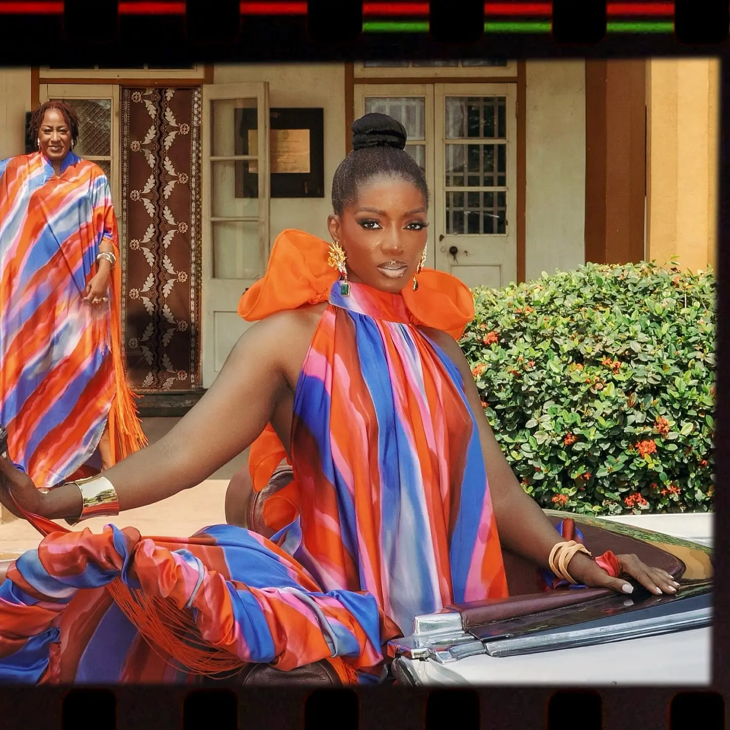 Model with a sleek bun in a striped orange-and-blue halter gown poses on a convertible outside a veranda.