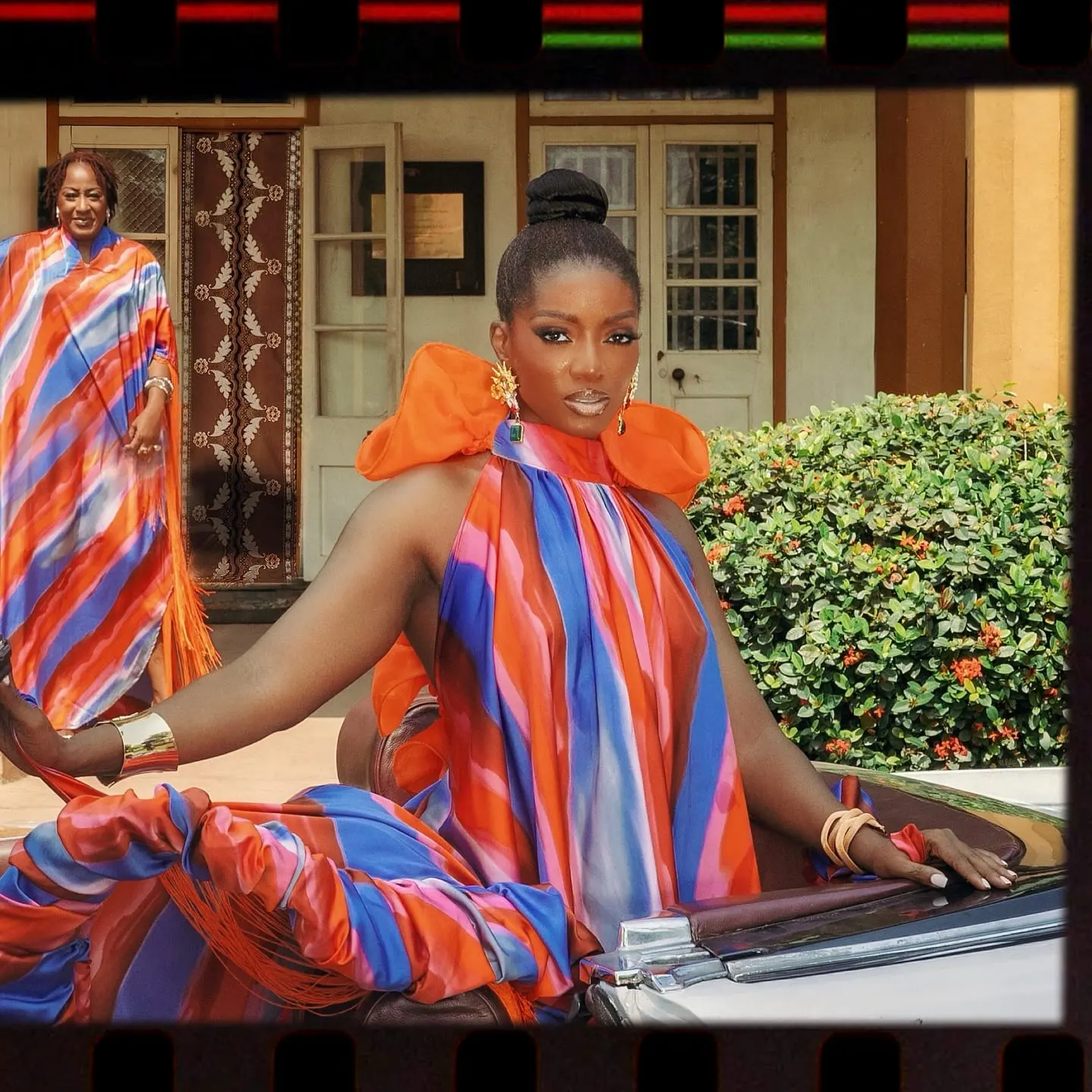 Model with a sleek bun in a striped orange-and-blue halter gown poses on a convertible outside a veranda.