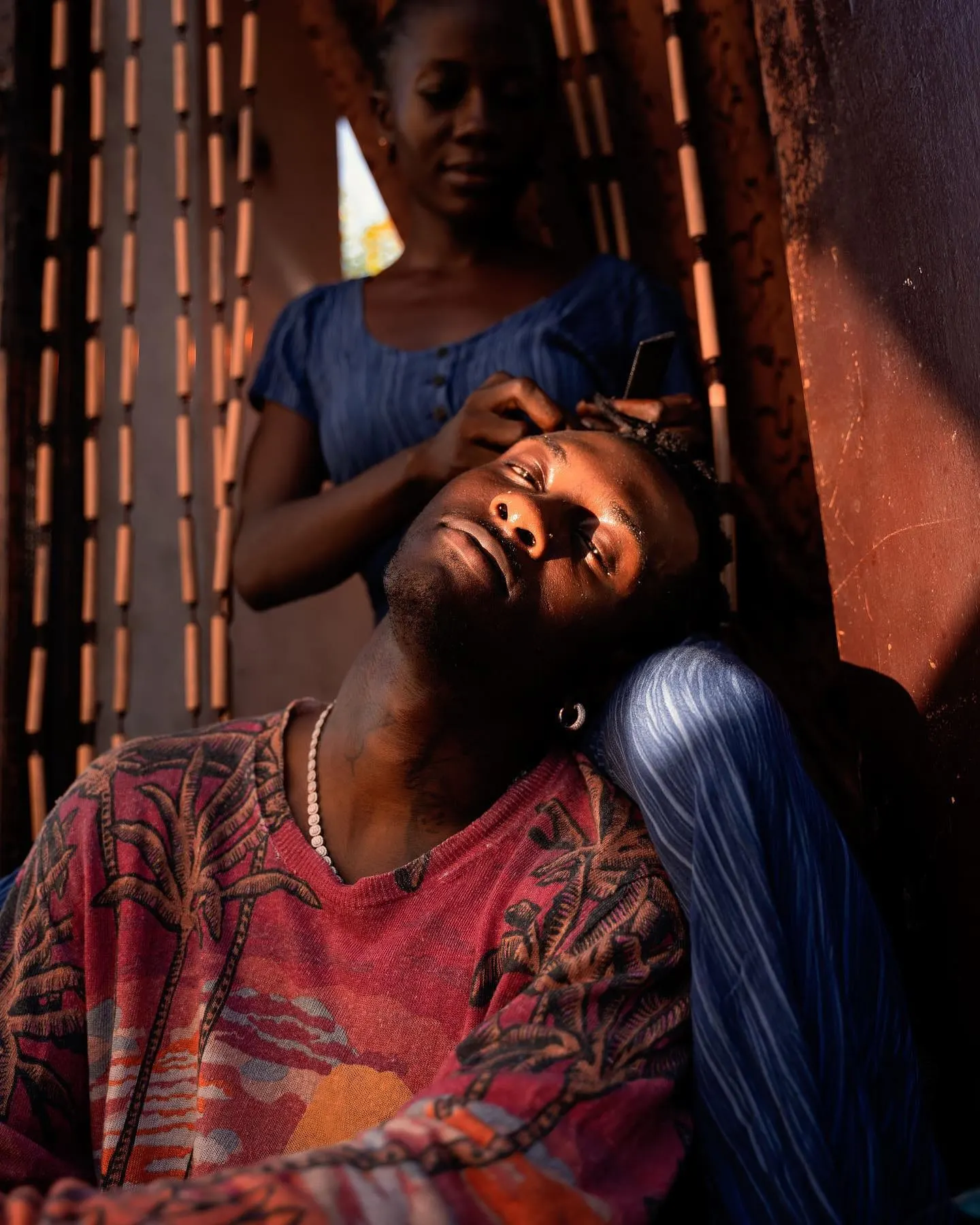 He rests his head back in warm evening light as a woman braids his hair in a doorway.