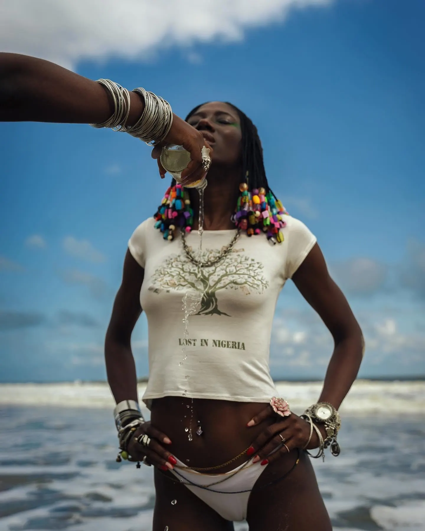 A drink is poured over a model in a Lost in Nigeria tee standing in the surf under blue sky.
