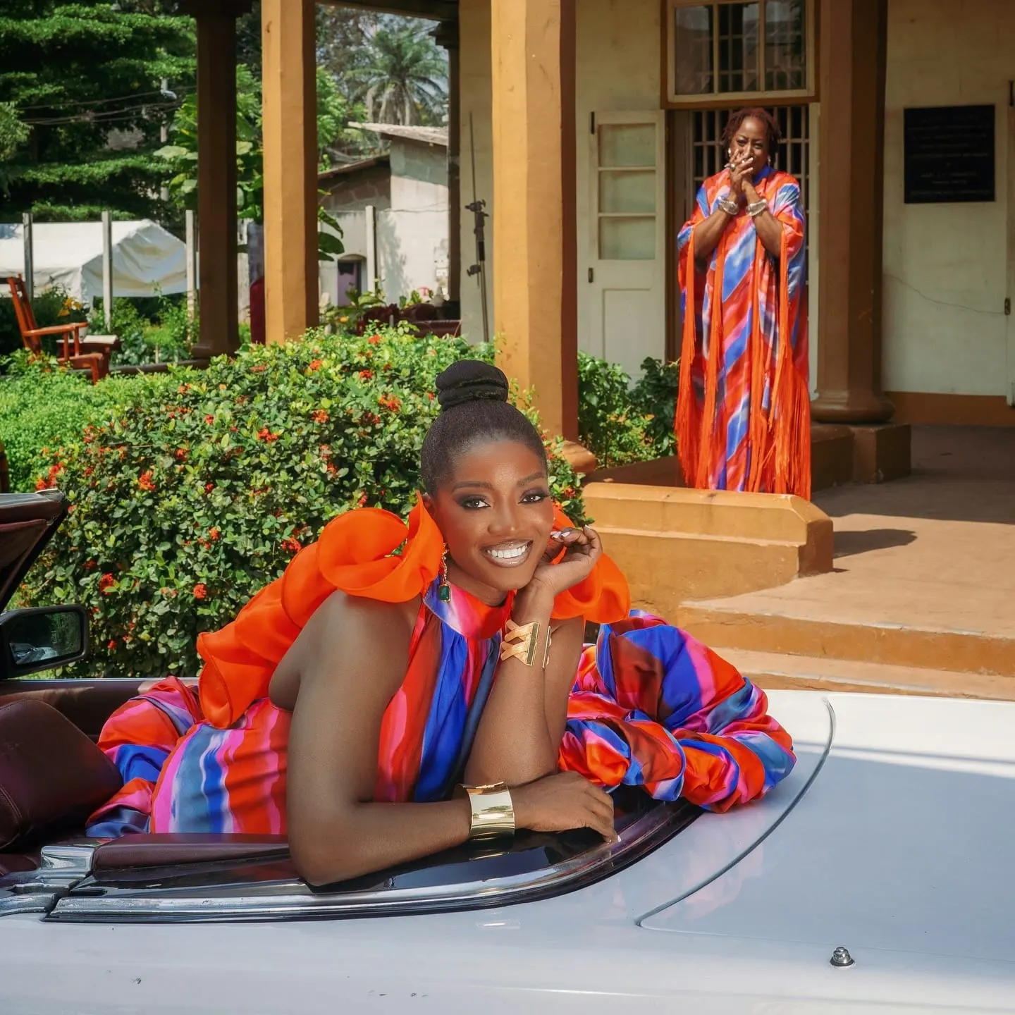Smiling model reclines on a white convertible in an orange-ruffled gown while an older woman applauds from the veranda.