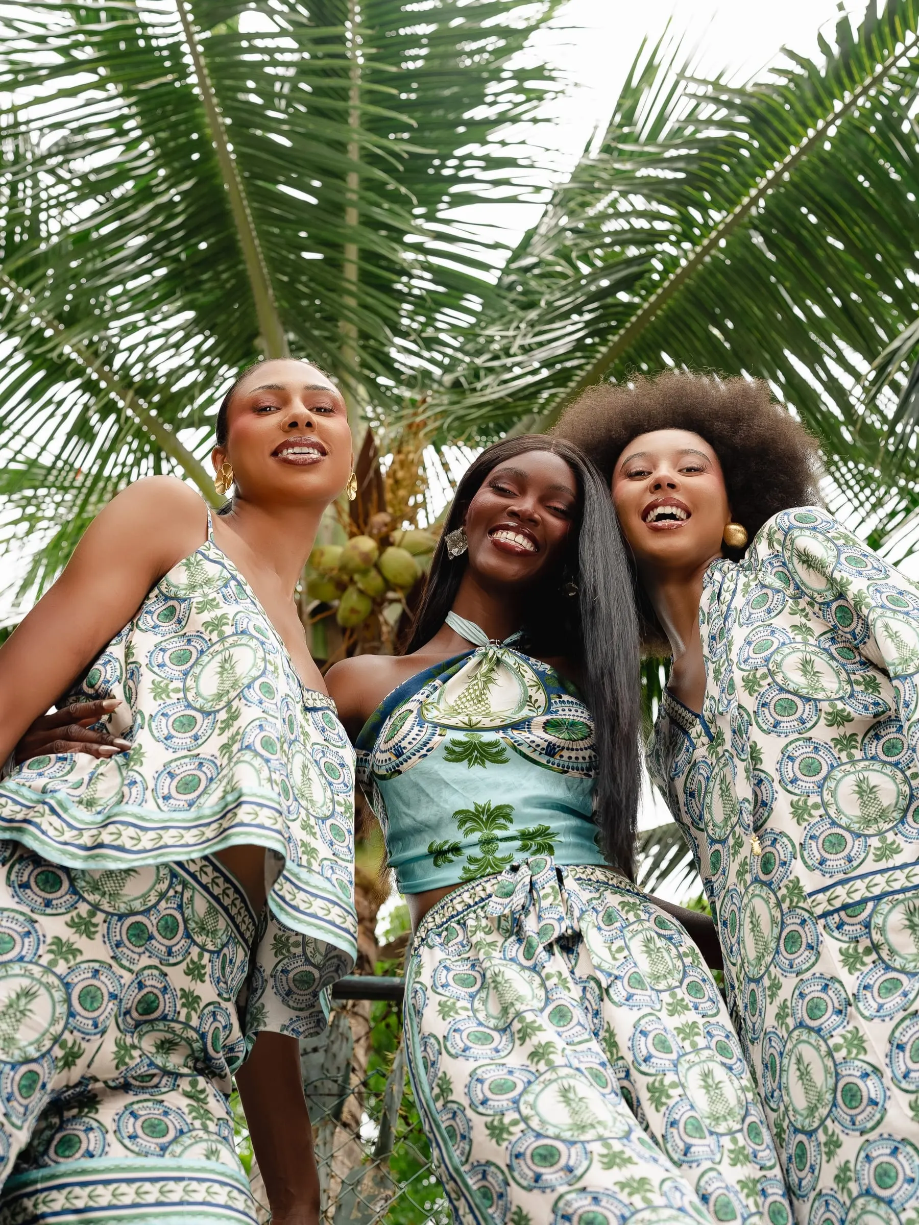 Three models laugh together beneath a coconut palm in matching blue-and-green paisley separates, photographed from below.