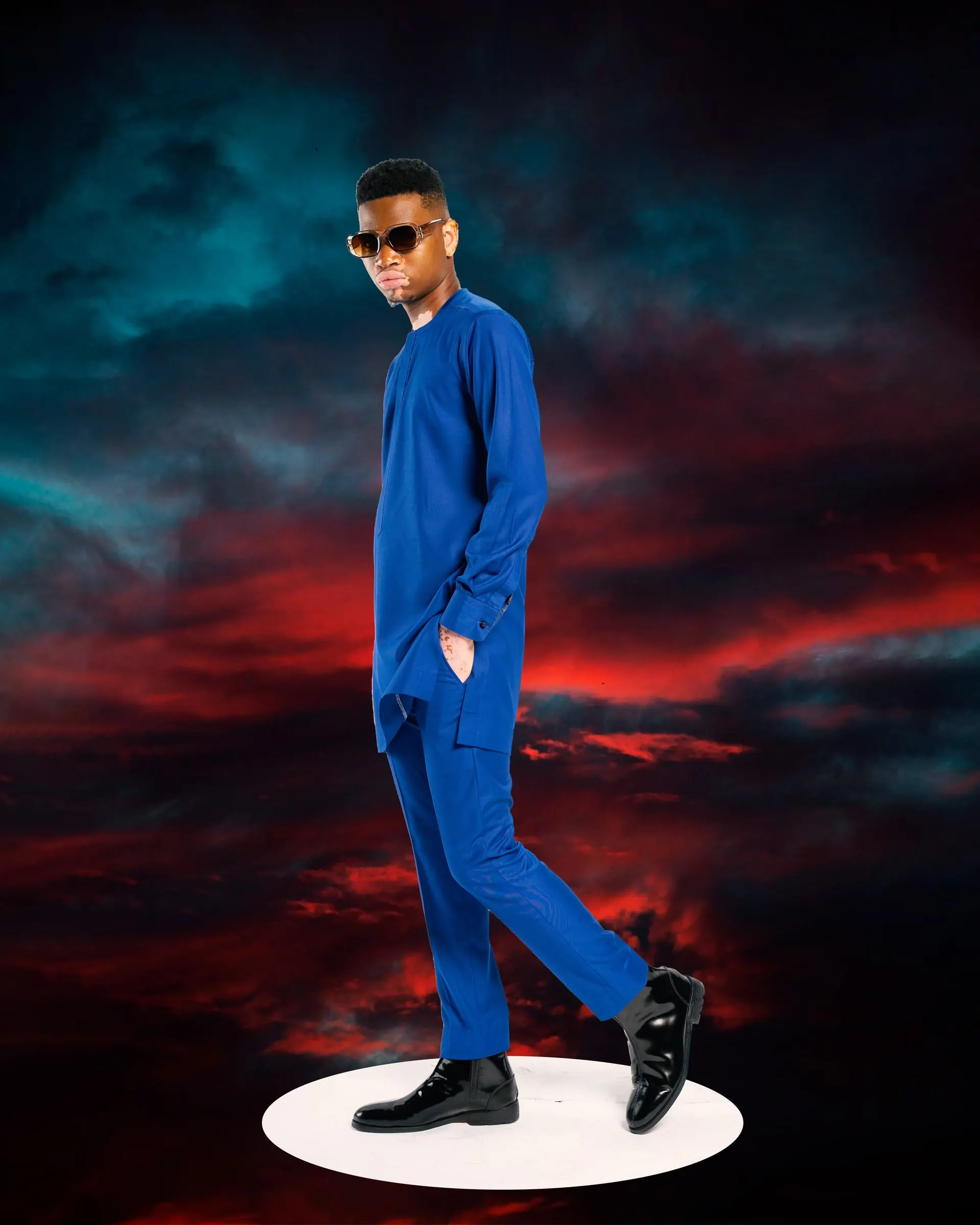 Model in a royal-blue tunic set and sunglasses strides across a white plinth, one hand in his pocket, against a storm sky.