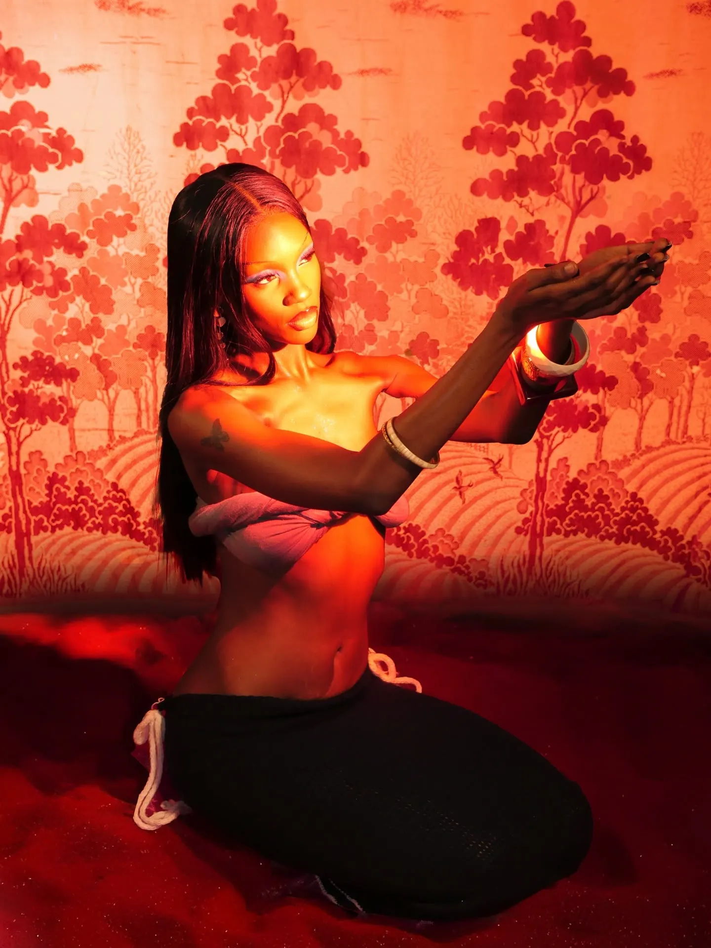 Kneeling model extends cupped hands toward warm light, half her face lit gold against the red toile set.