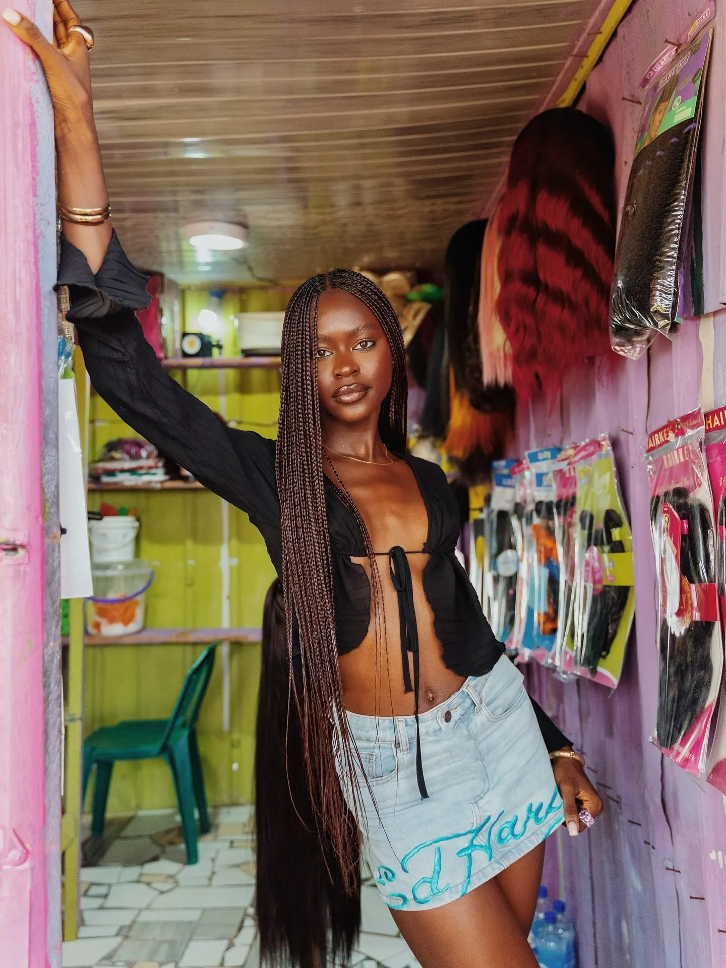 Model with floor-length brown braids posing in a pink kiosk doorway lined with packaged hair extensions.