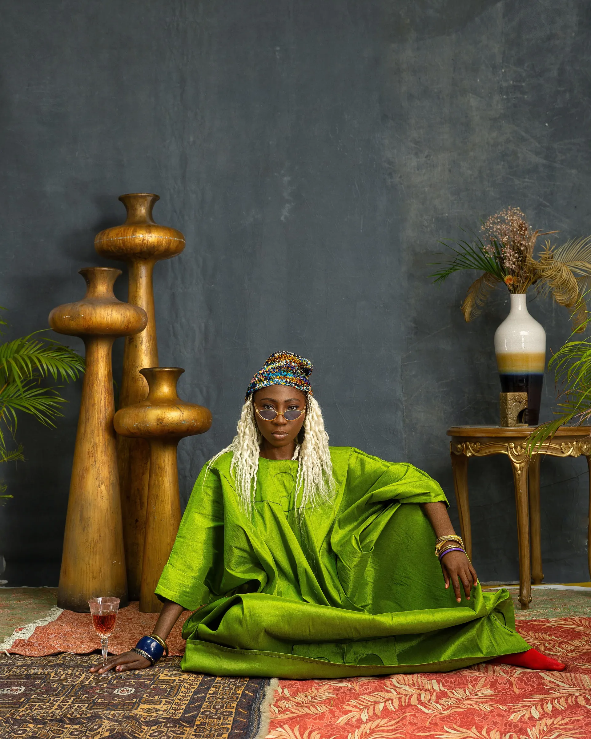 Model with long white braids, a beaded cap and round glasses sits on layered rugs in a lime-green boubou, one knee raised, a stemmed glass on the rug beside tall gold vessels.