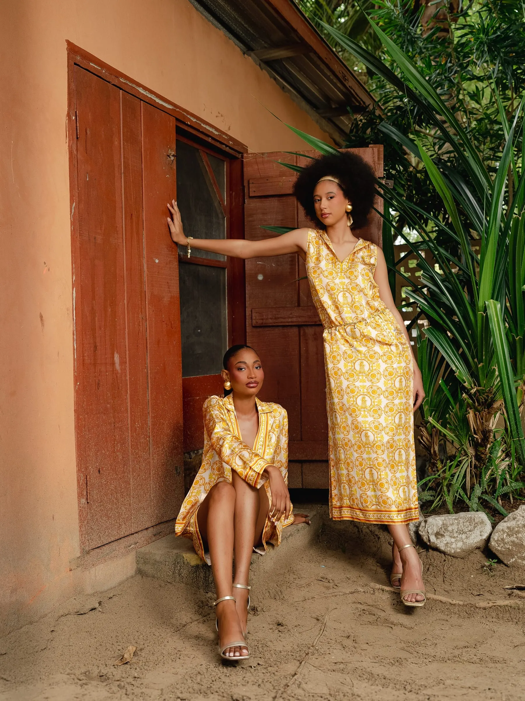 Two models at a rust-red wooden door: one seated on the step in a yellow printed shirt dress, the other standing in a sleeveless yellow patterned midi dress with one arm on the frame.
