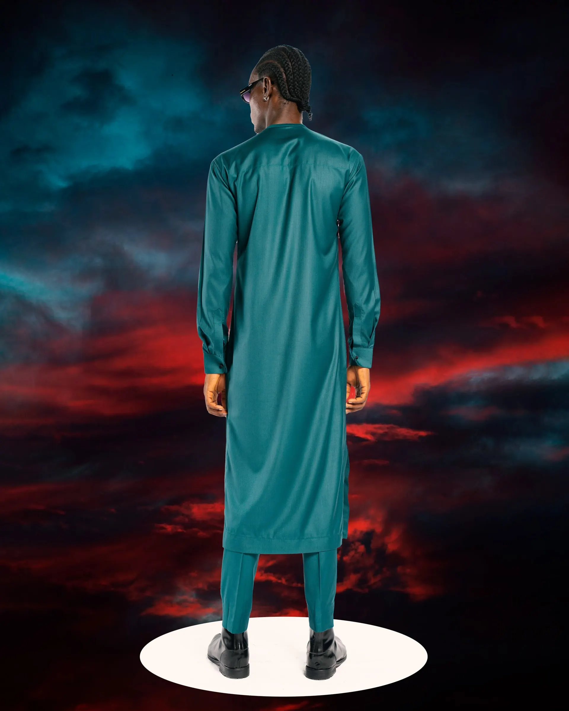 Model seen from behind in a long teal kaftan on a white plinth, looking back over his shoulder at a storm sky.