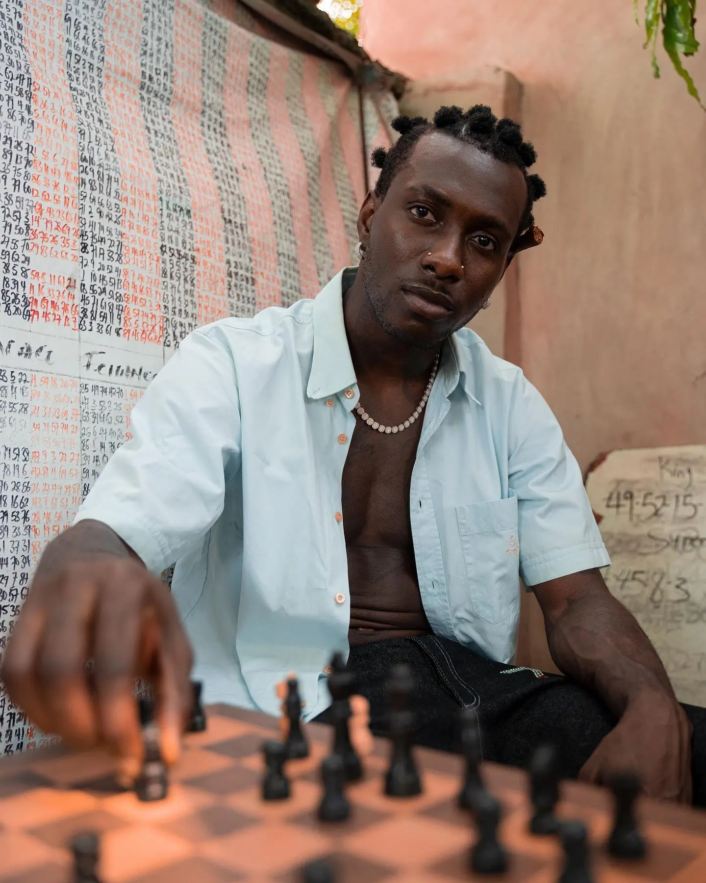 Wearing bantu knots and a pale blue shirt, he moves a chess piece before a numbers-covered wall.