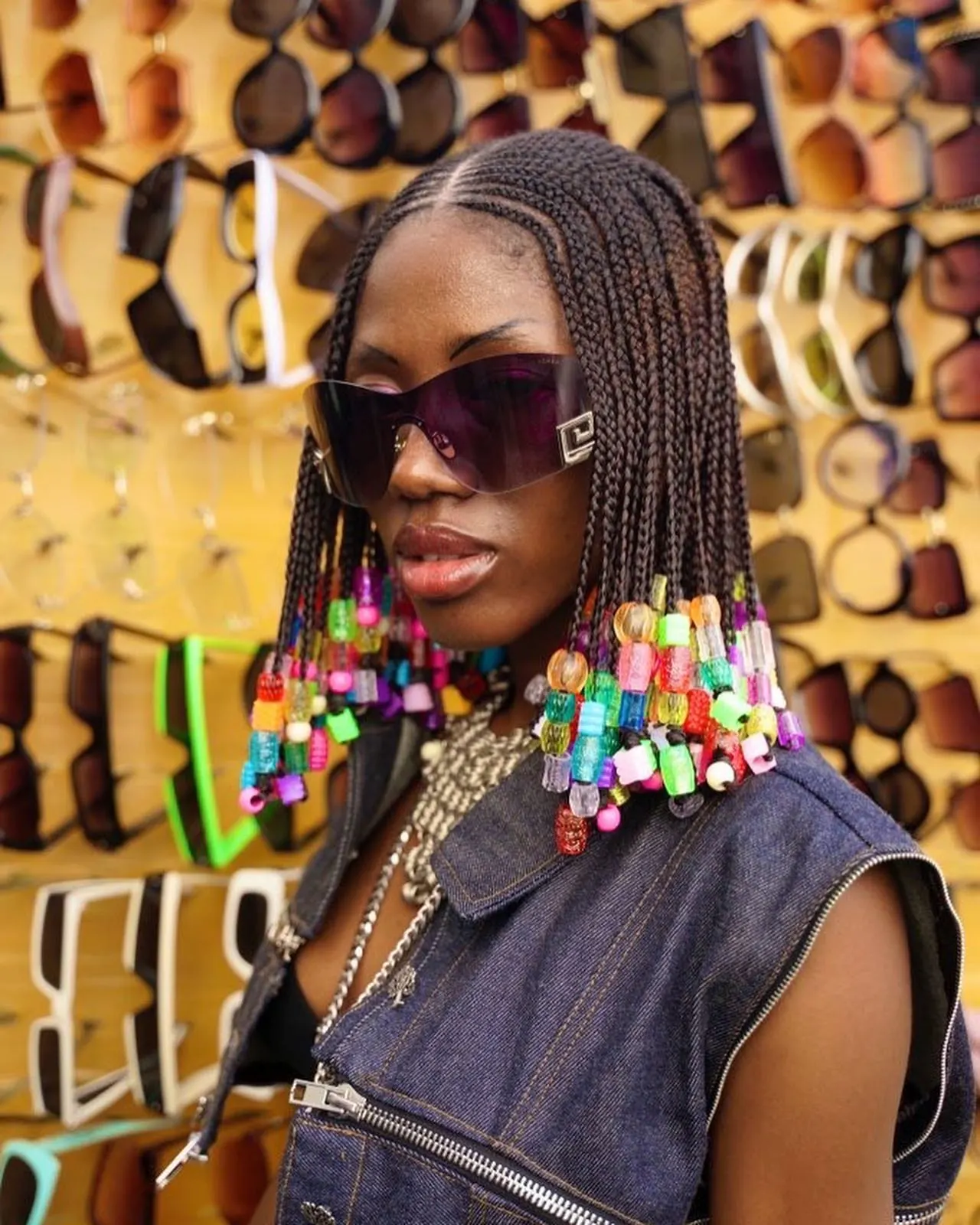 Close portrait of a model in tinted shield sunglasses, braids ending in rainbow beads, before a wall of sunglasses.
