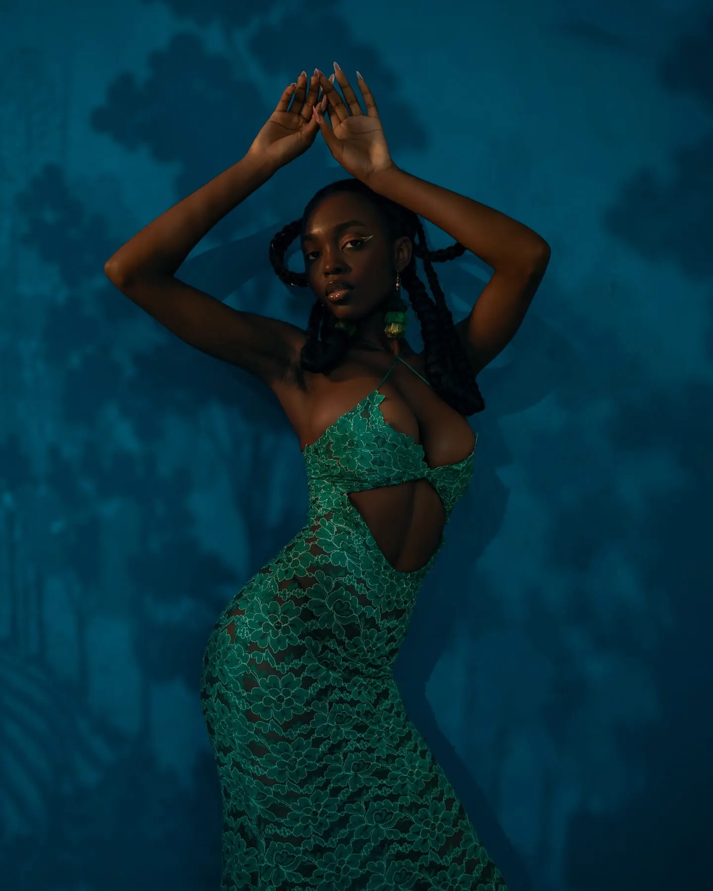 Uyi arches with arms raised overhead, the cutout green lace gown catching warm light against blue shadows.
