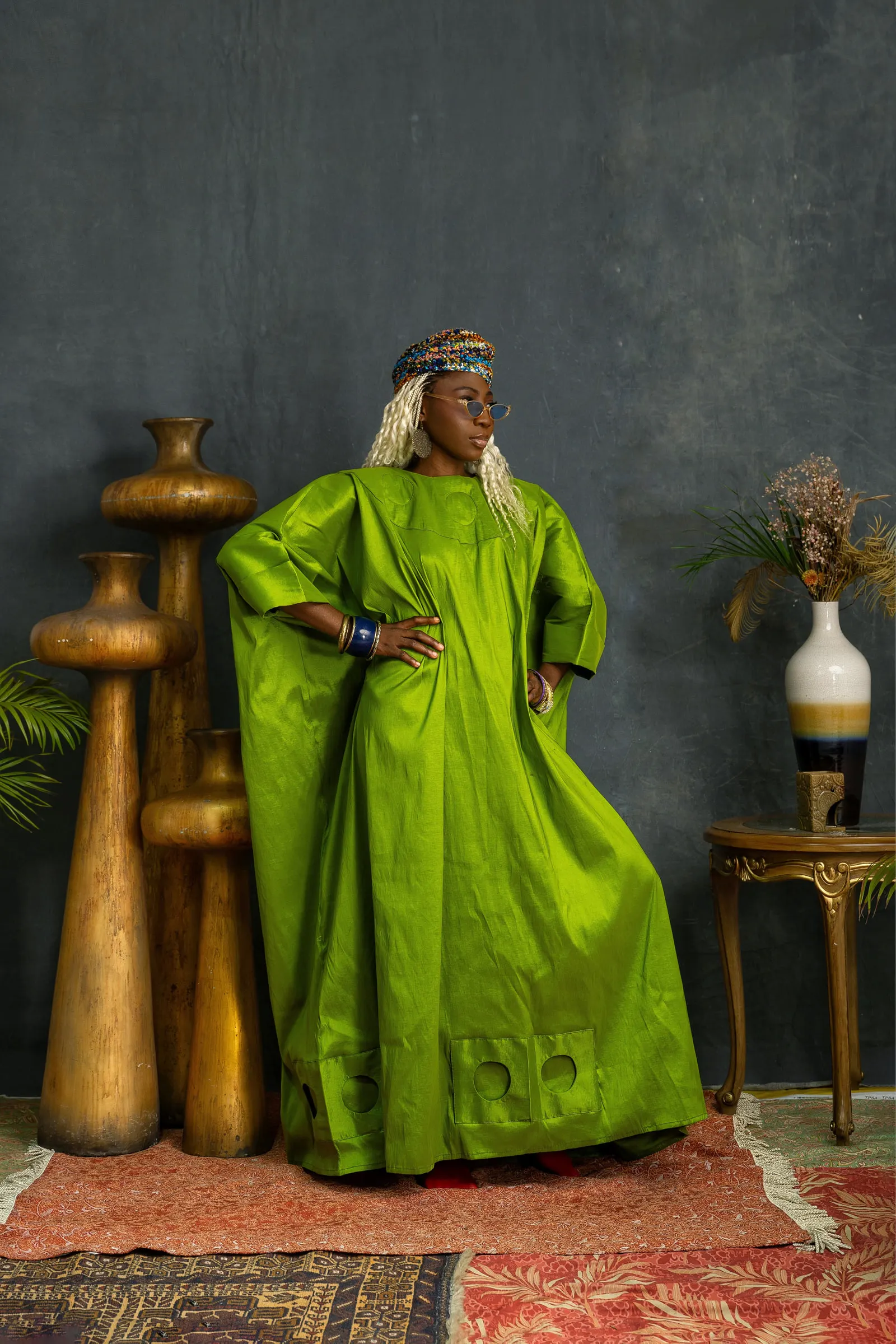 Model with white braids and a beaded cap stands with one hand on the hip in a lime-green boubou with circular motifs along the hem, gold vessels and a vase of dried grasses behind.