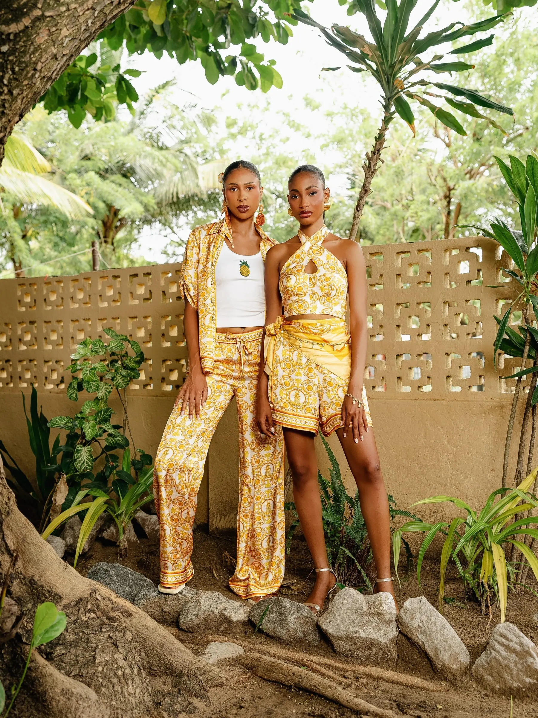 Two models in golden-yellow printed separates, a pineapple motif on a white vest, stand before a breeze-block wall under palms.