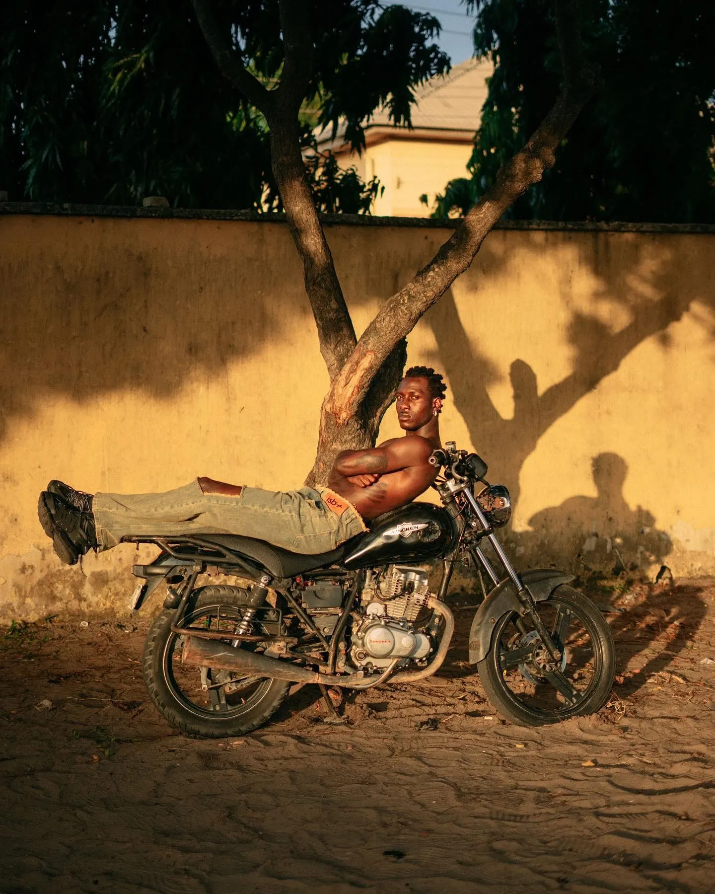 Reclining shirtless along a motorcycle seat beneath a tree, legs stretched out against a yellow wall.