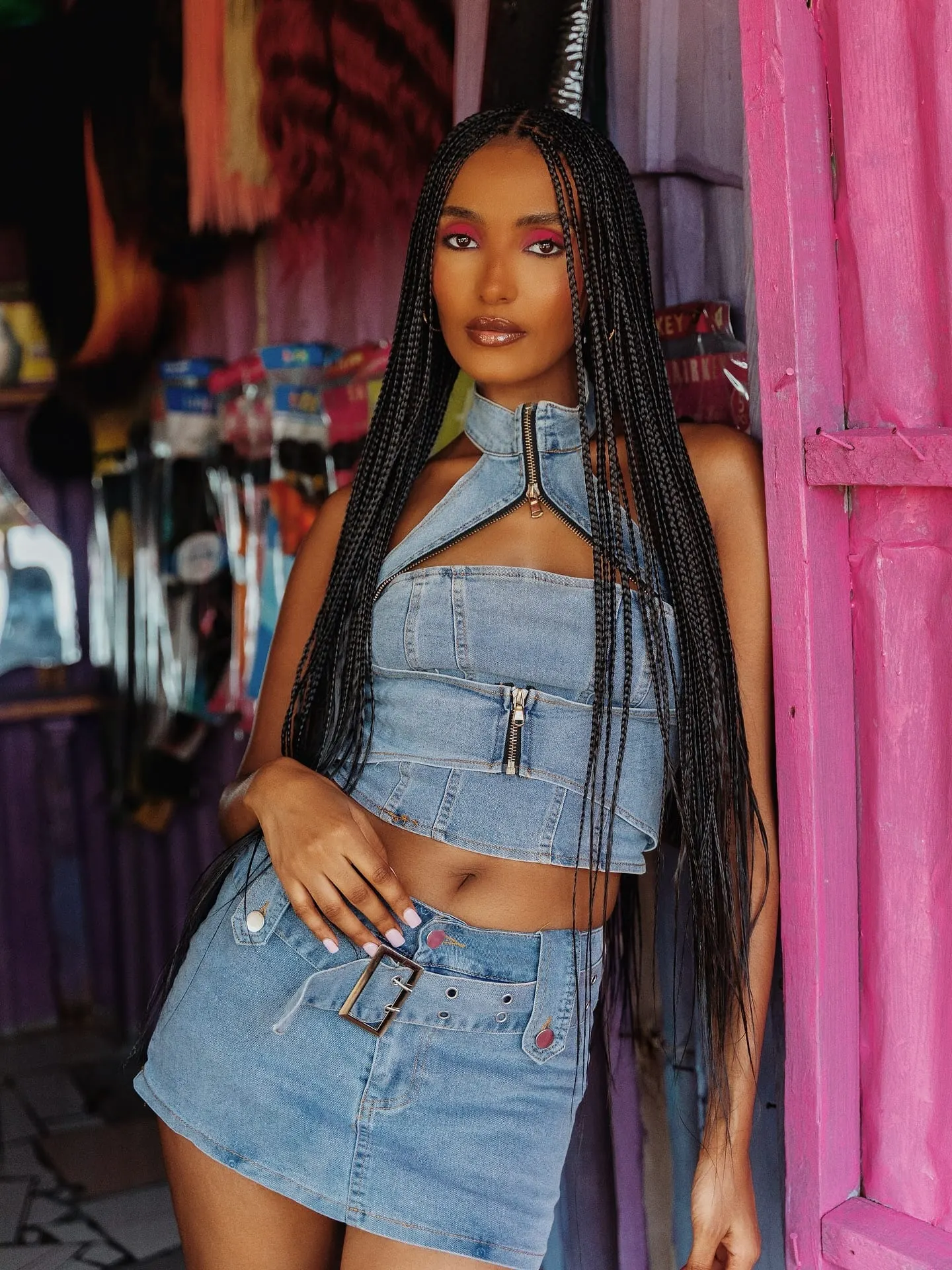 Model with long black braids and pink eyeshadow, in a zip-detail denim set, leaning on a pink kiosk wall.