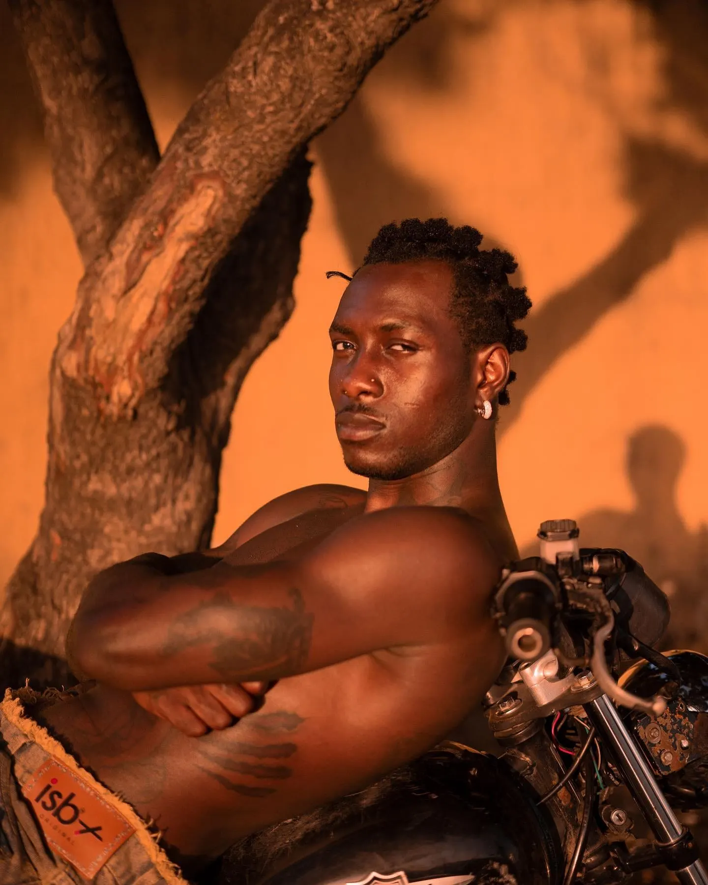 Close portrait leaning on motorcycle handlebars at sunset, tattooed arms crossed, bantu knots catching amber light.