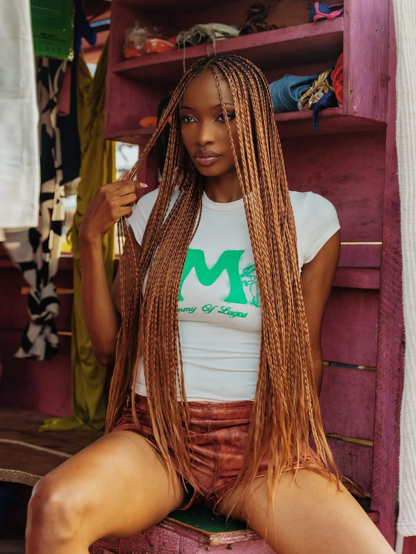 Model holding a strand of her copper braids, seated in a pink stall wearing a Mummy of Lagos tee.