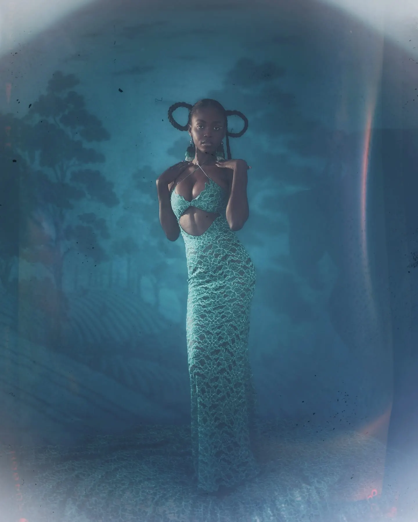 Full-length film frame of Uyi standing in soft haze, hands at her collarbone, braids looped like a crown.