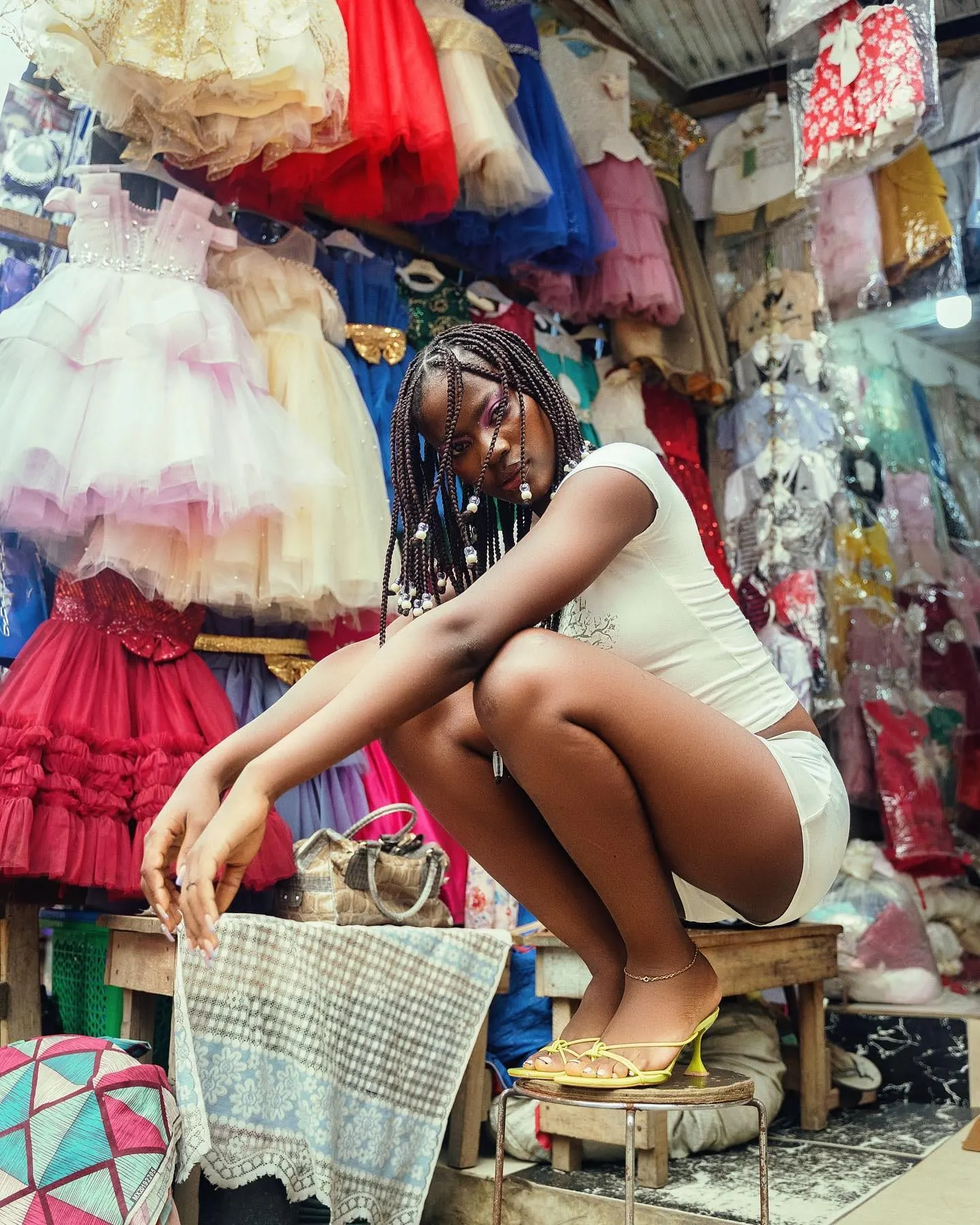 Model in white crop top and yellow heels perches on a stool amid racks of frilly tulle dresses.