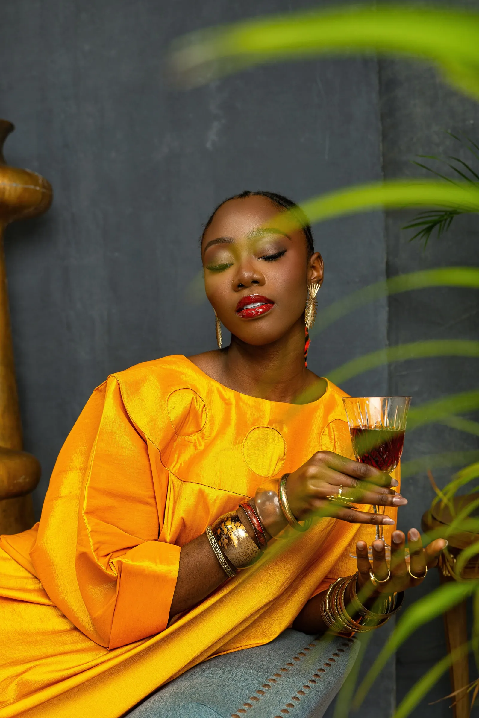 Model in a marigold-orange boubou reclines in a pale armchair with eyes closed, raising a stemmed glass, palm fronds blurred across the foreground.
