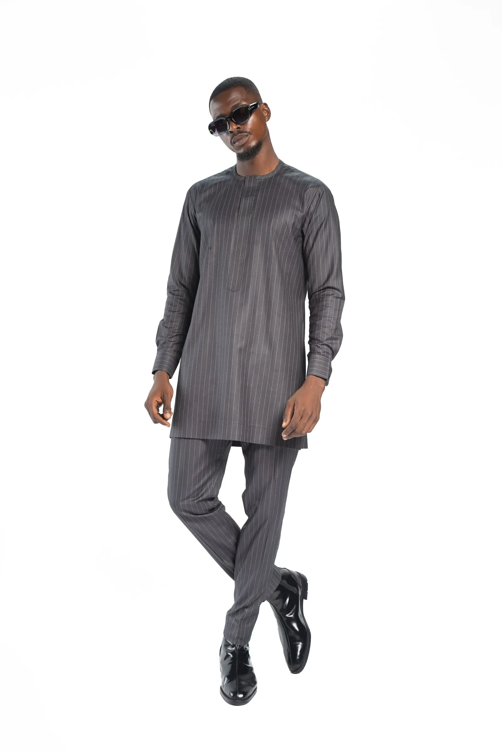 Model in a grey pinstripe tunic set and sunglasses crosses one leg over the other against a white studio background.