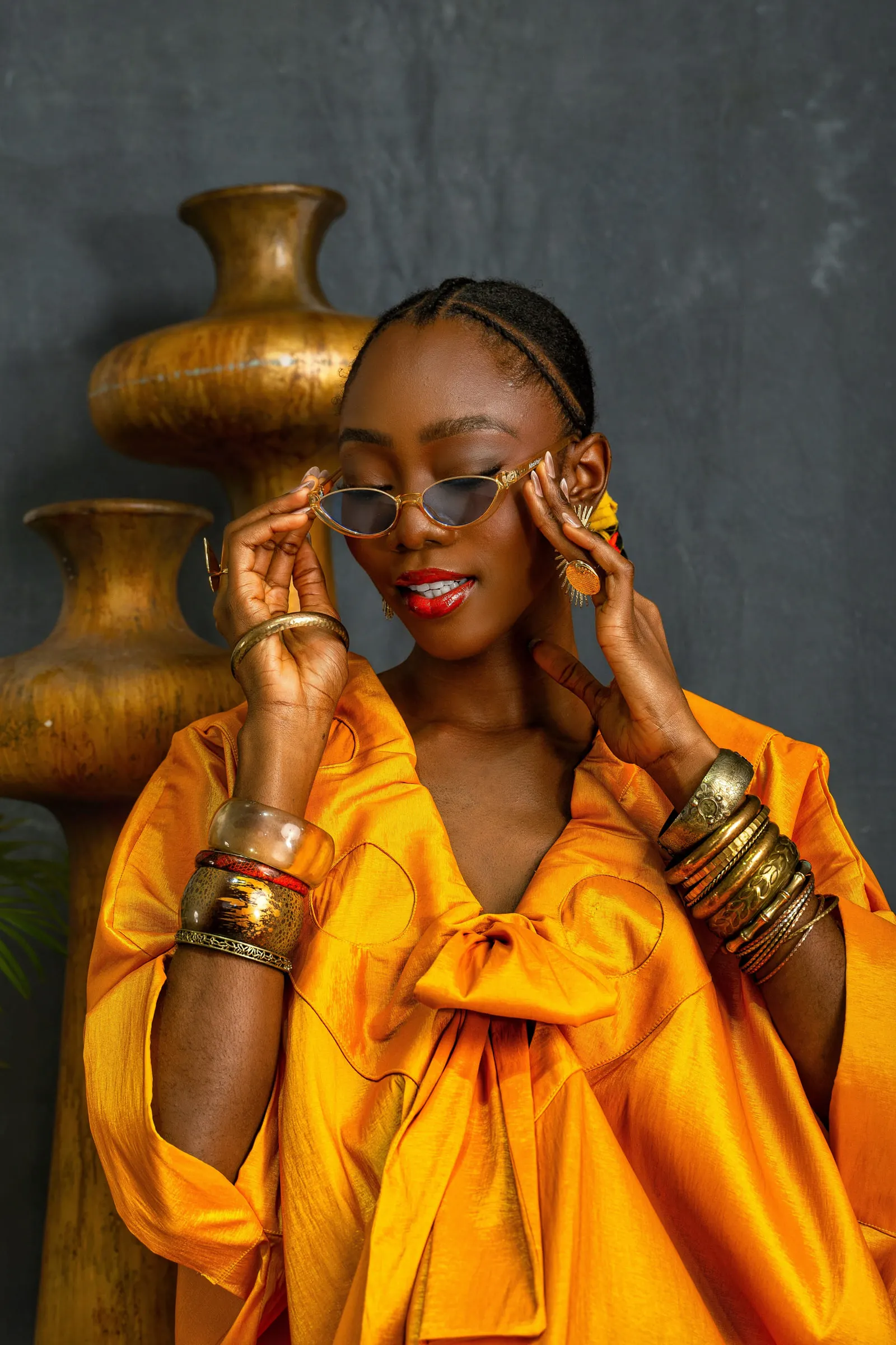 Close portrait of a model in a marigold-orange boubou lifting slim sunglasses with both hands, stacked gold and red bangles on each wrist, a gold vessel behind.