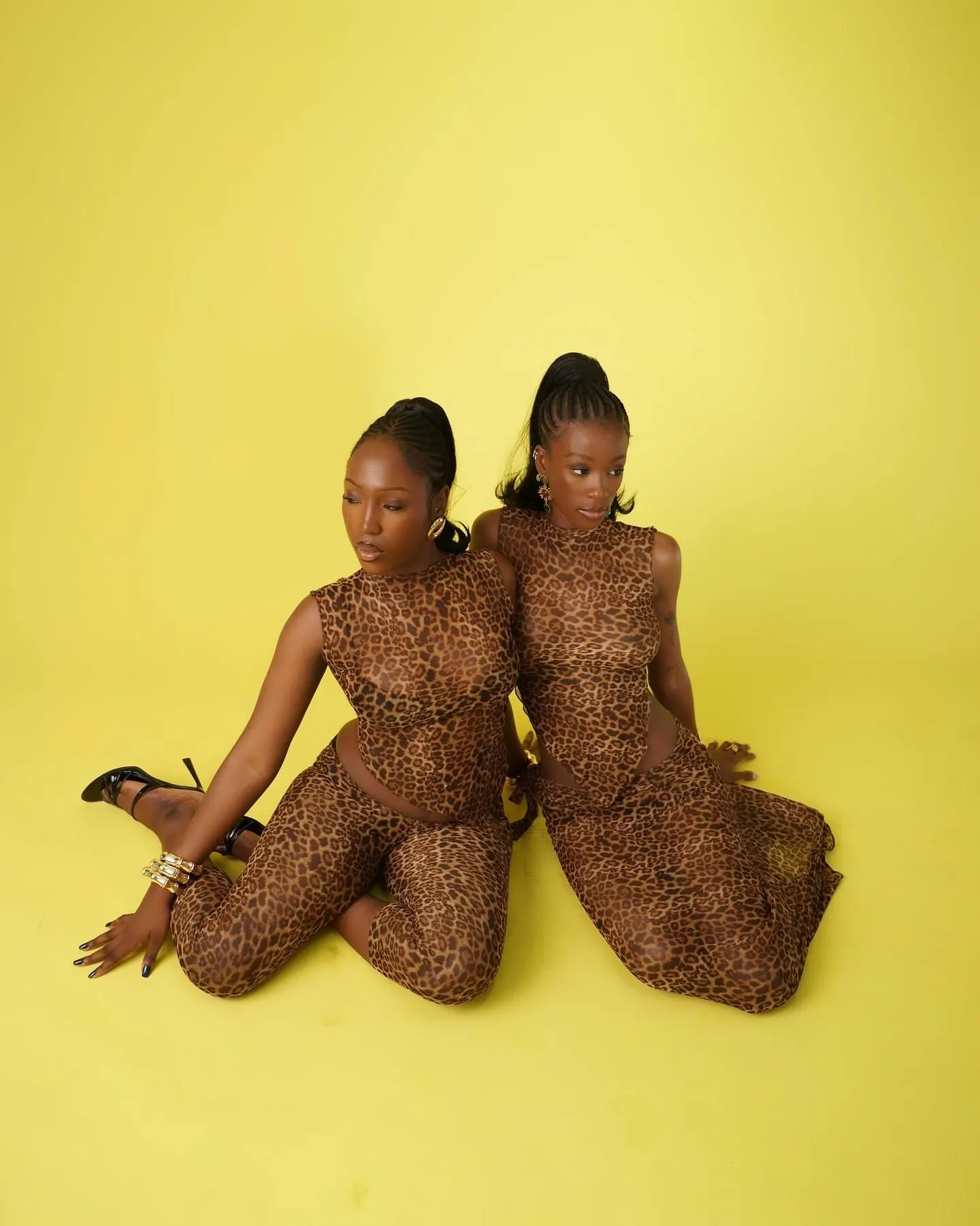 Two models in matching sheer leopard-print sets sitting back to back on a yellow studio floor.