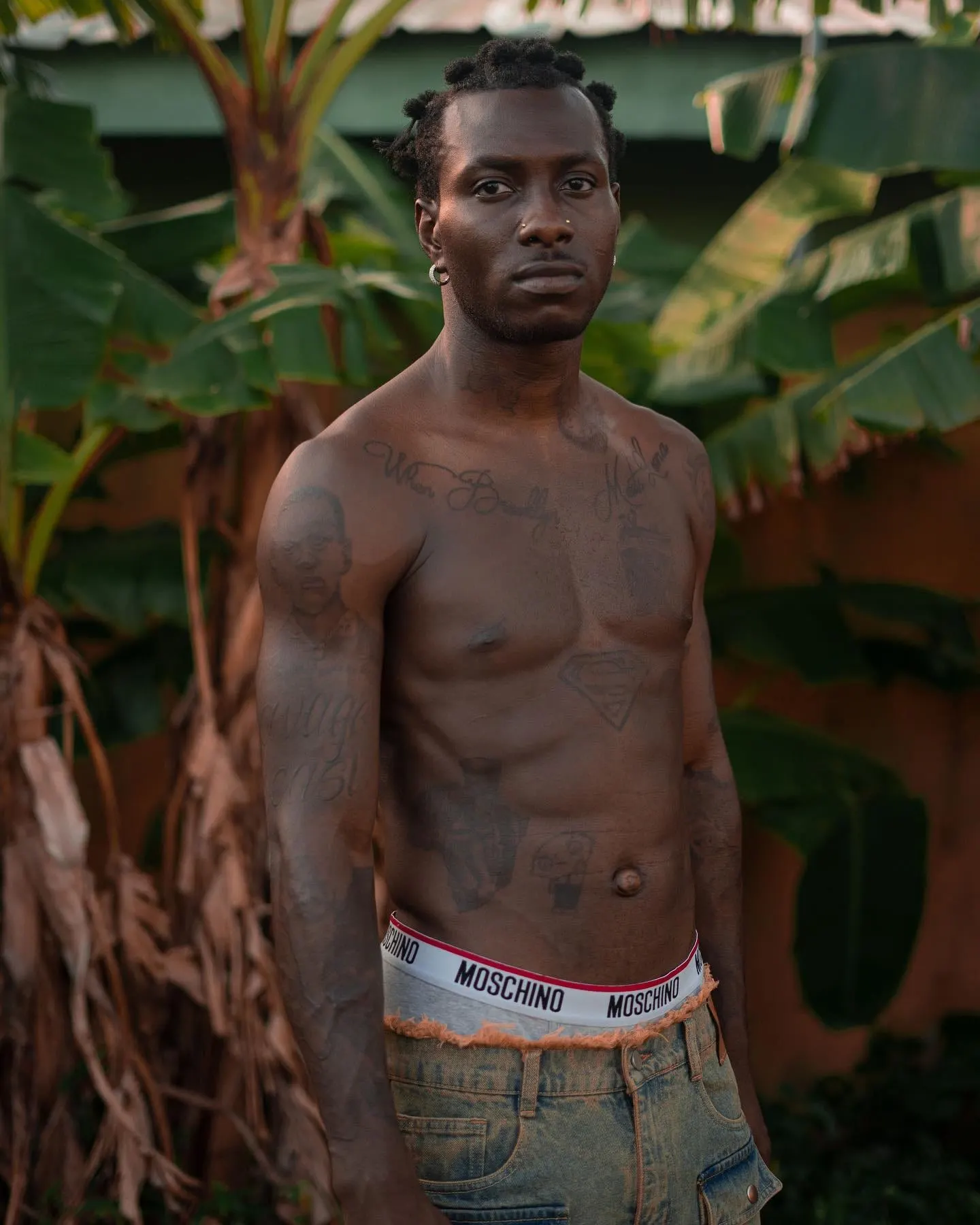 Shirtless portrait among banana leaves, tattoos across his chest and arms, Moschino waistband above faded jeans.
