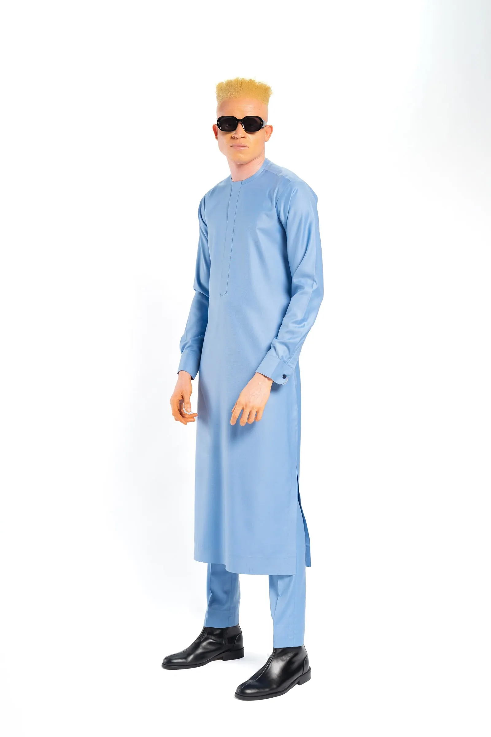 Model with a blond flat-top in a long light-blue kaftan and sunglasses stands in profile against a white background.