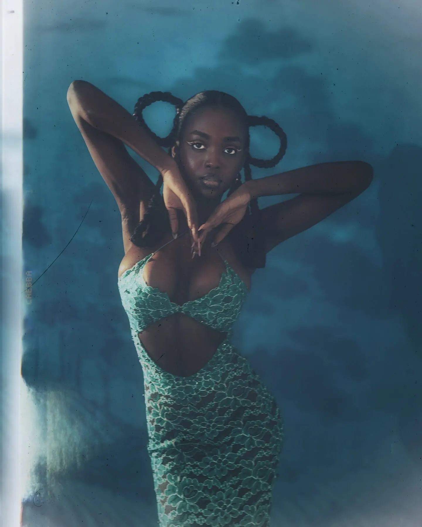 Film still of Uyi facing the camera, arms bent behind her head, dark skin glowing against misty blue.
