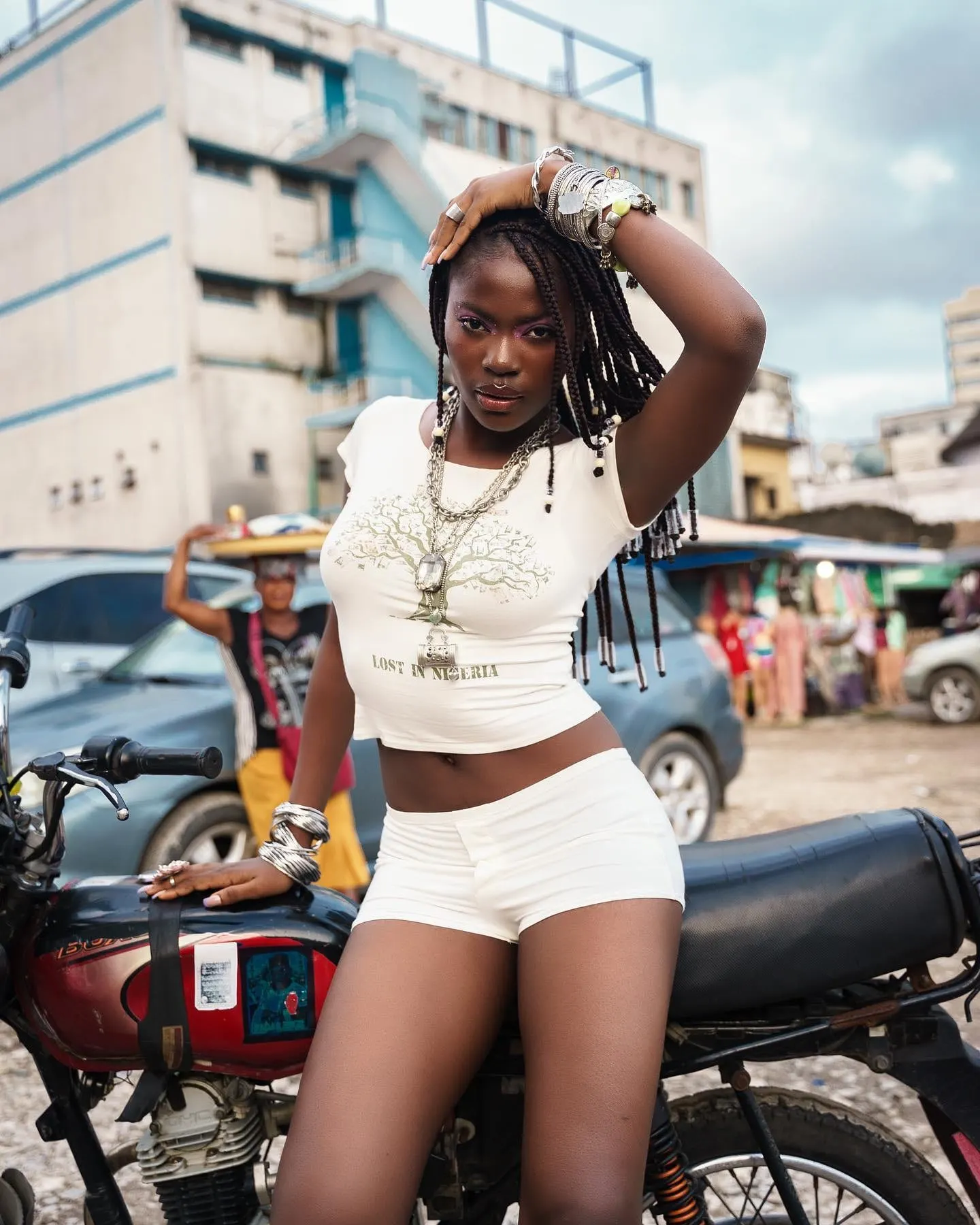 Model in a Lost in Nigeria tee and white shorts leans on a motorcycle, hand on her braids.