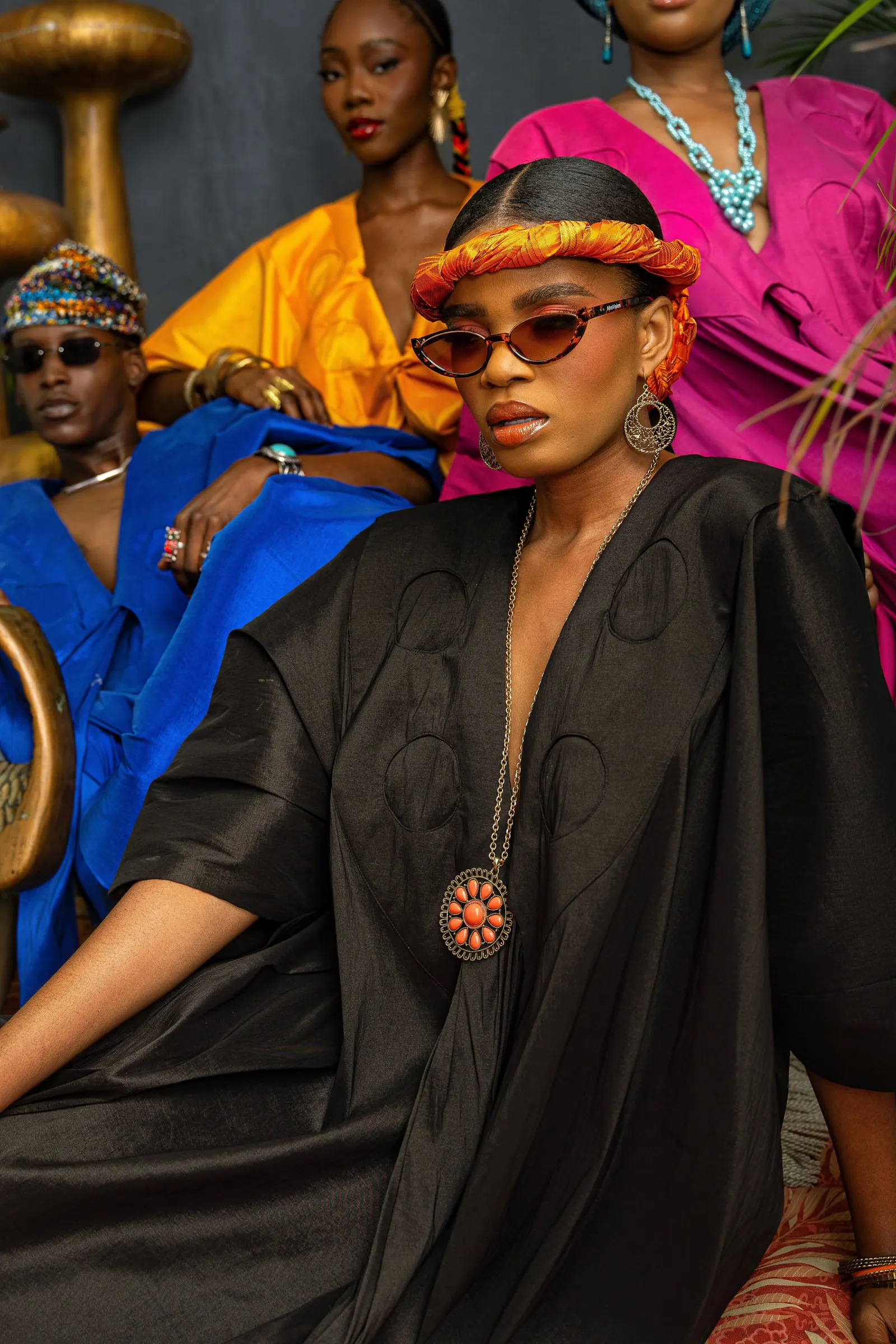 Model in a black boubou with an orange braided headband, tinted sunglasses and an orange stone pendant sits in front, with models in blue, orange and pink out of focus behind.