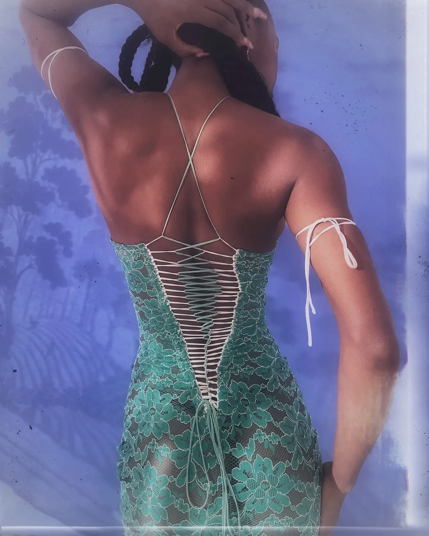 Back view of the gown's corset lacing, Uyi's hands lifted into her braids against a lilac-blue wall.